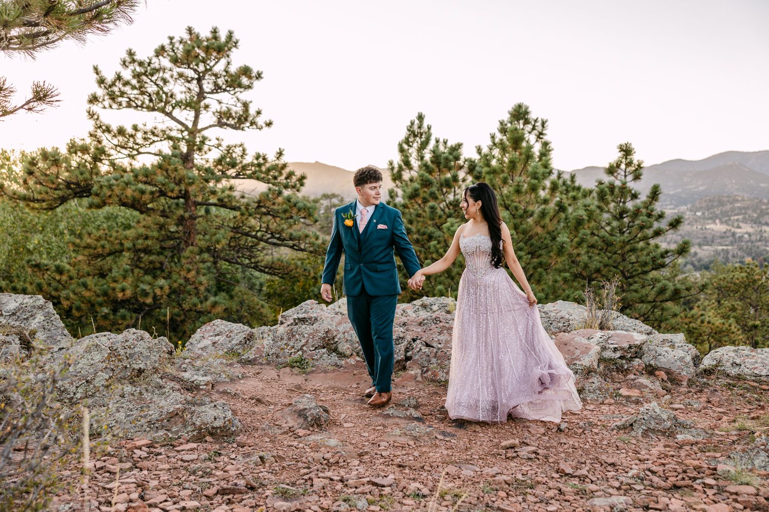 A couple in formal attire walks hand in hand along a rocky mountain overlook with pine trees and distant hills.