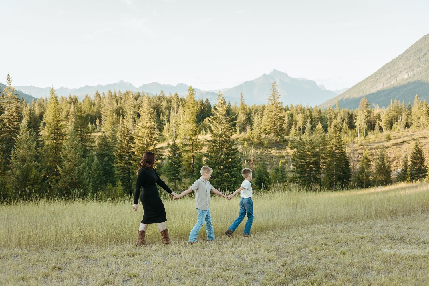 People in blue dresses and black outfits dance joyfully in a golden mountain meadow at sunset with evergreen trees behind them.