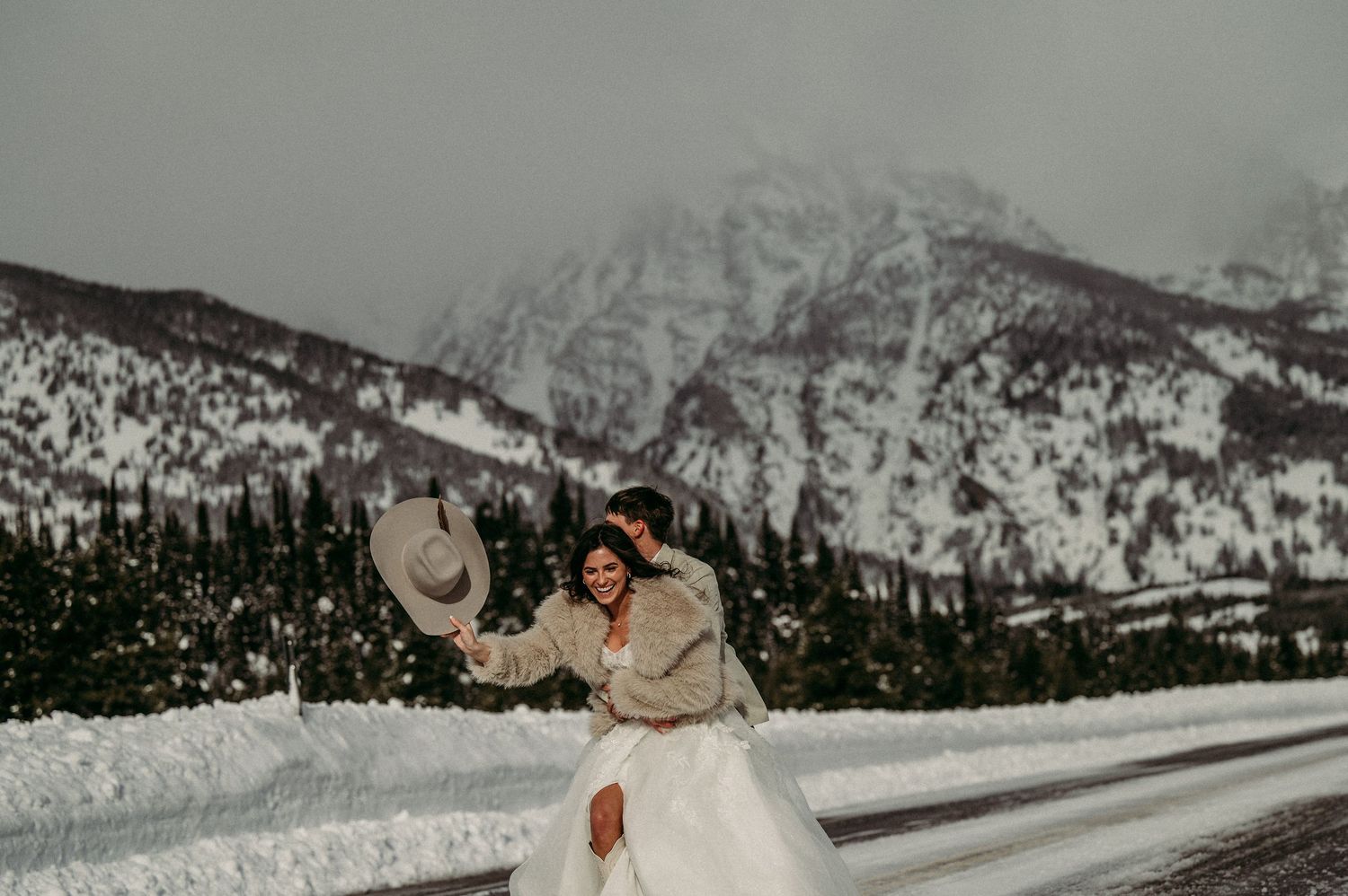 Bride and groom embrace on snowy mountain road in winter wedding attire, with snow-covered peaks in the background.