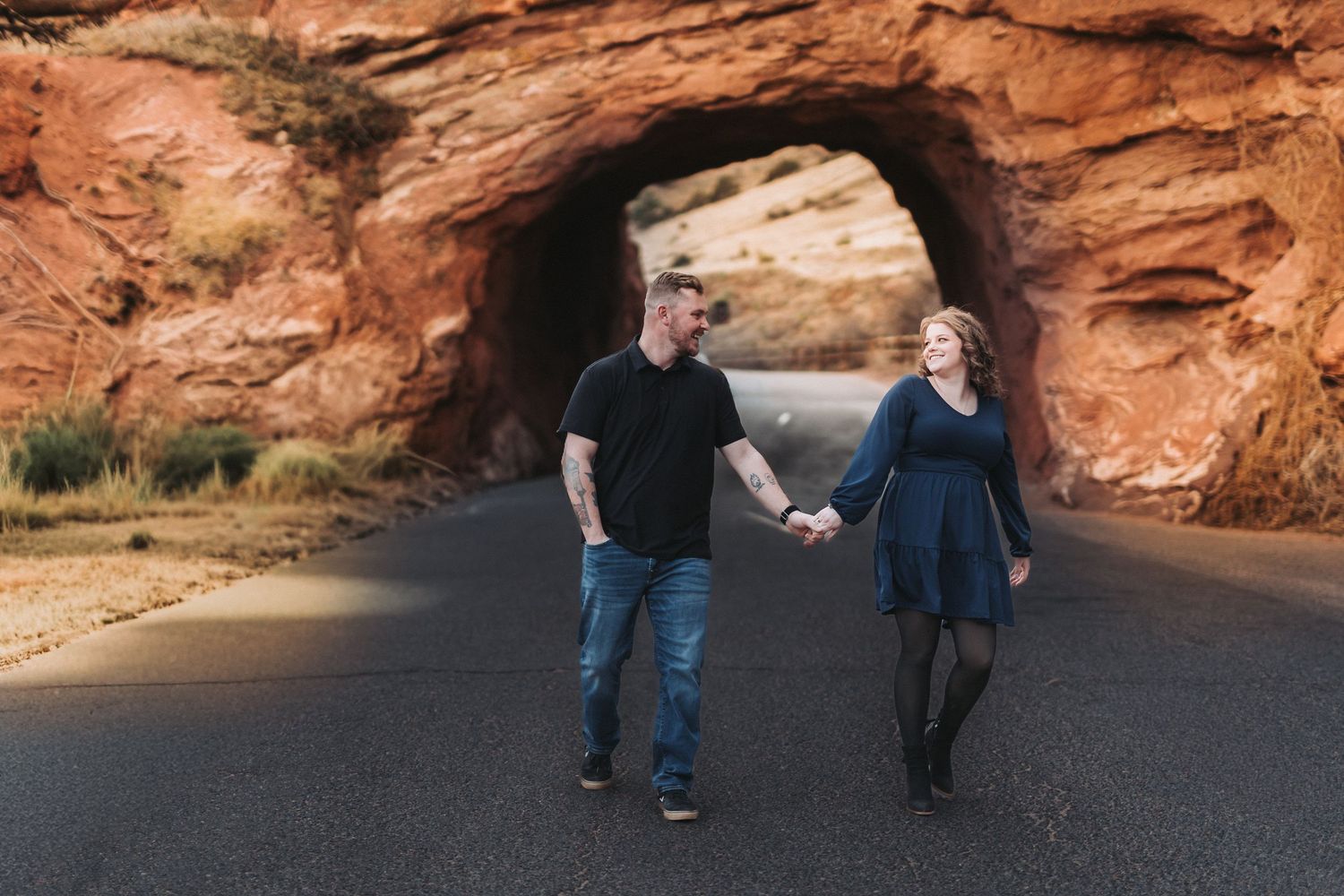 Couple walking hand in hand through a red rock tunnel formation on a paved road.