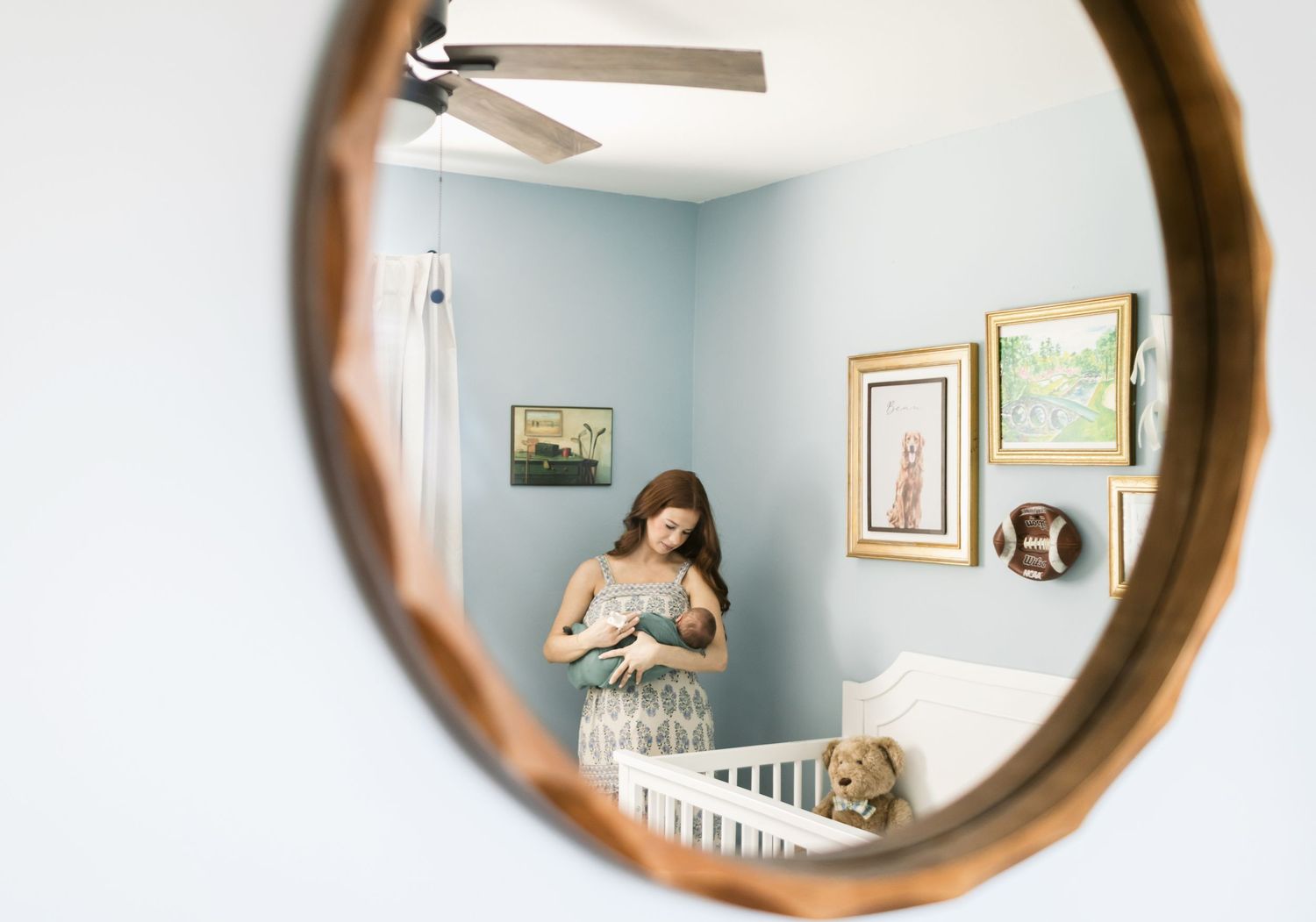 Mother with red hair stands in corner of the baby nursery with blue walls holding her newborn swaddled in seafood green for newborn photos