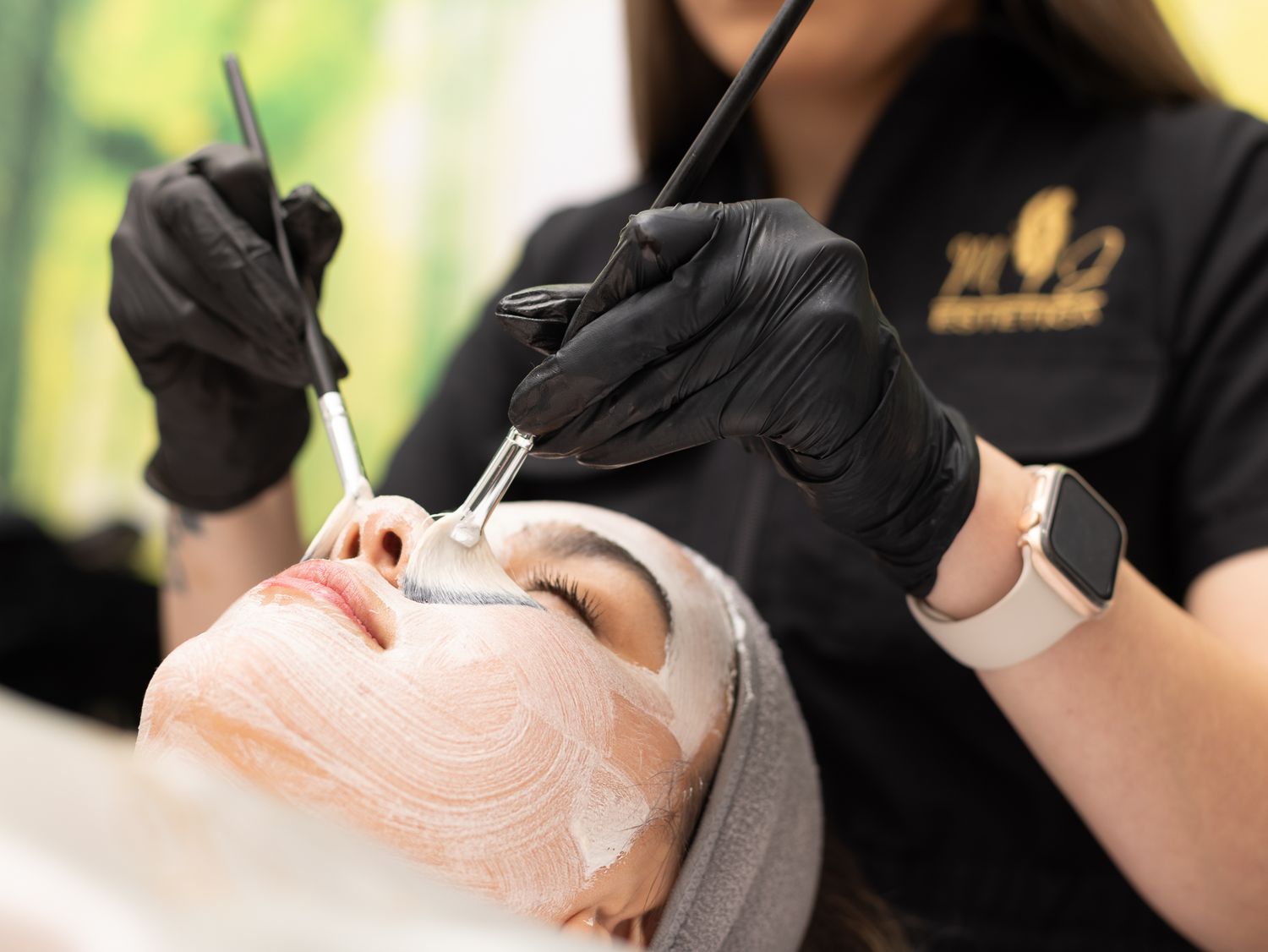 Esthetician in black gloves applying a facial mask treatment to a client at a professional beauty spa.