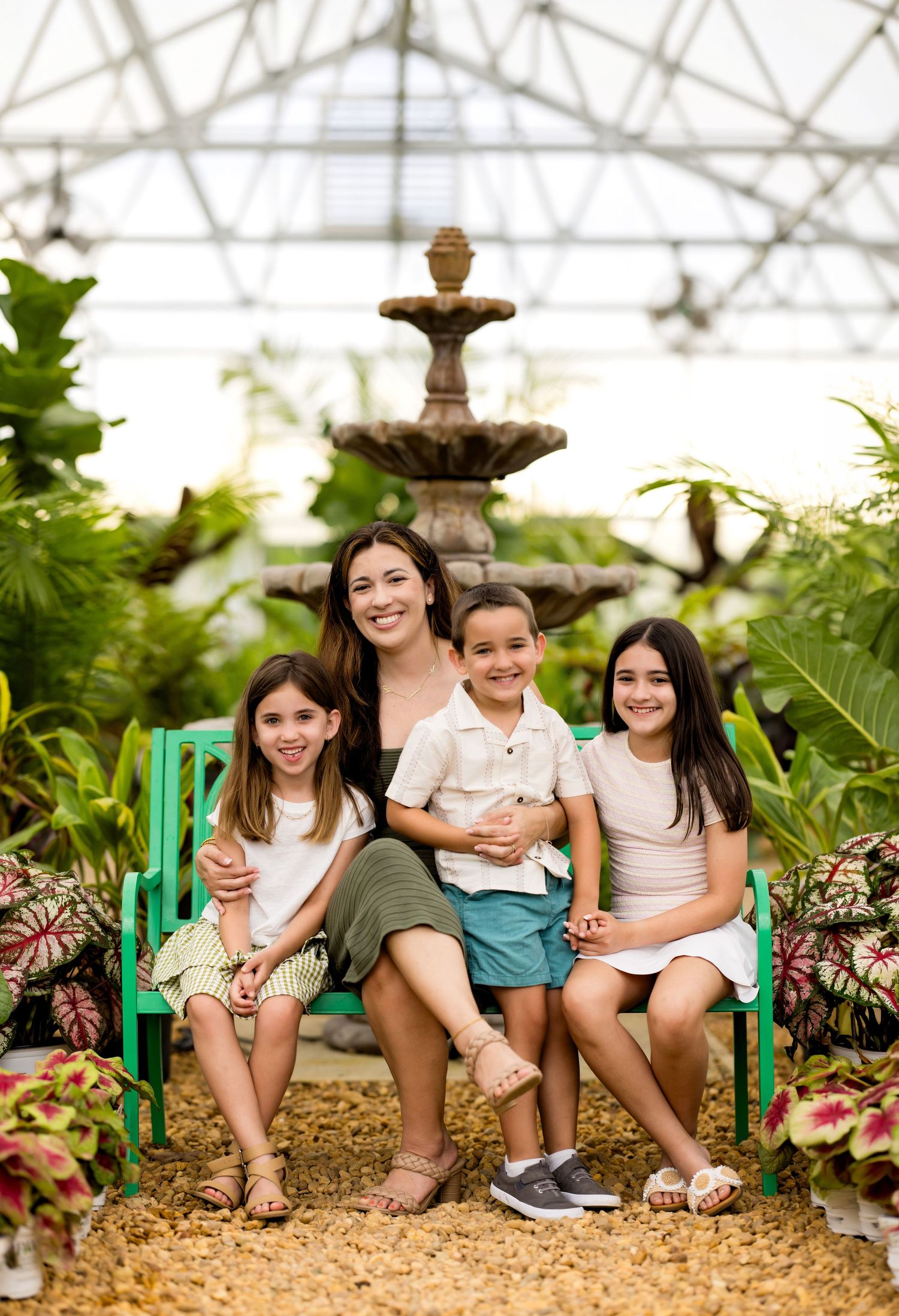 A smiling woman and three children pose on a green bench in a lush greenhouse with a stone fountain backdrop.