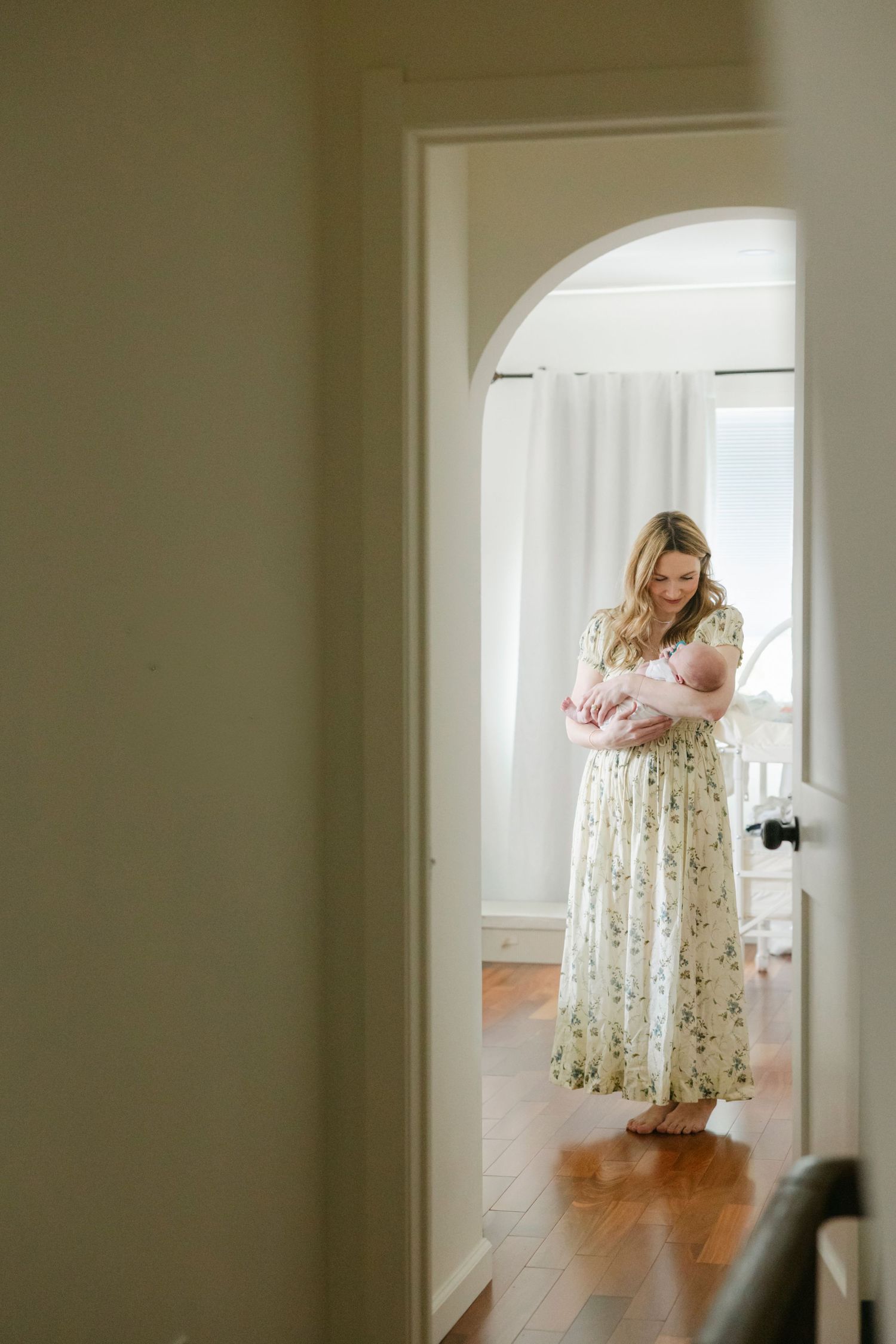 A mother in a floral dress holds her newborn baby in a sunlit room framed by an elegant arched doorway.