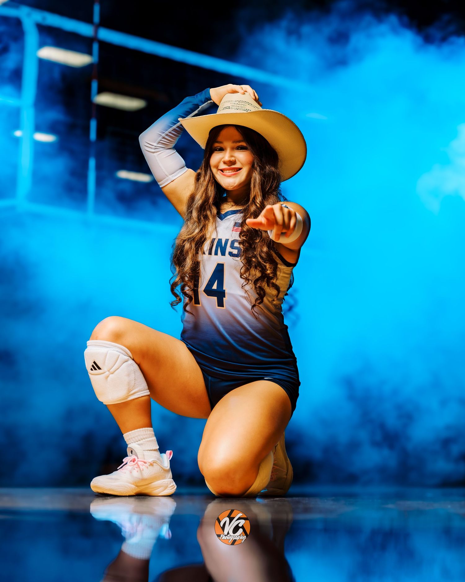 Female volleyball player #4 poses in blue jersey and cowboy hat amid dramatic blue smoke on a reflective court.