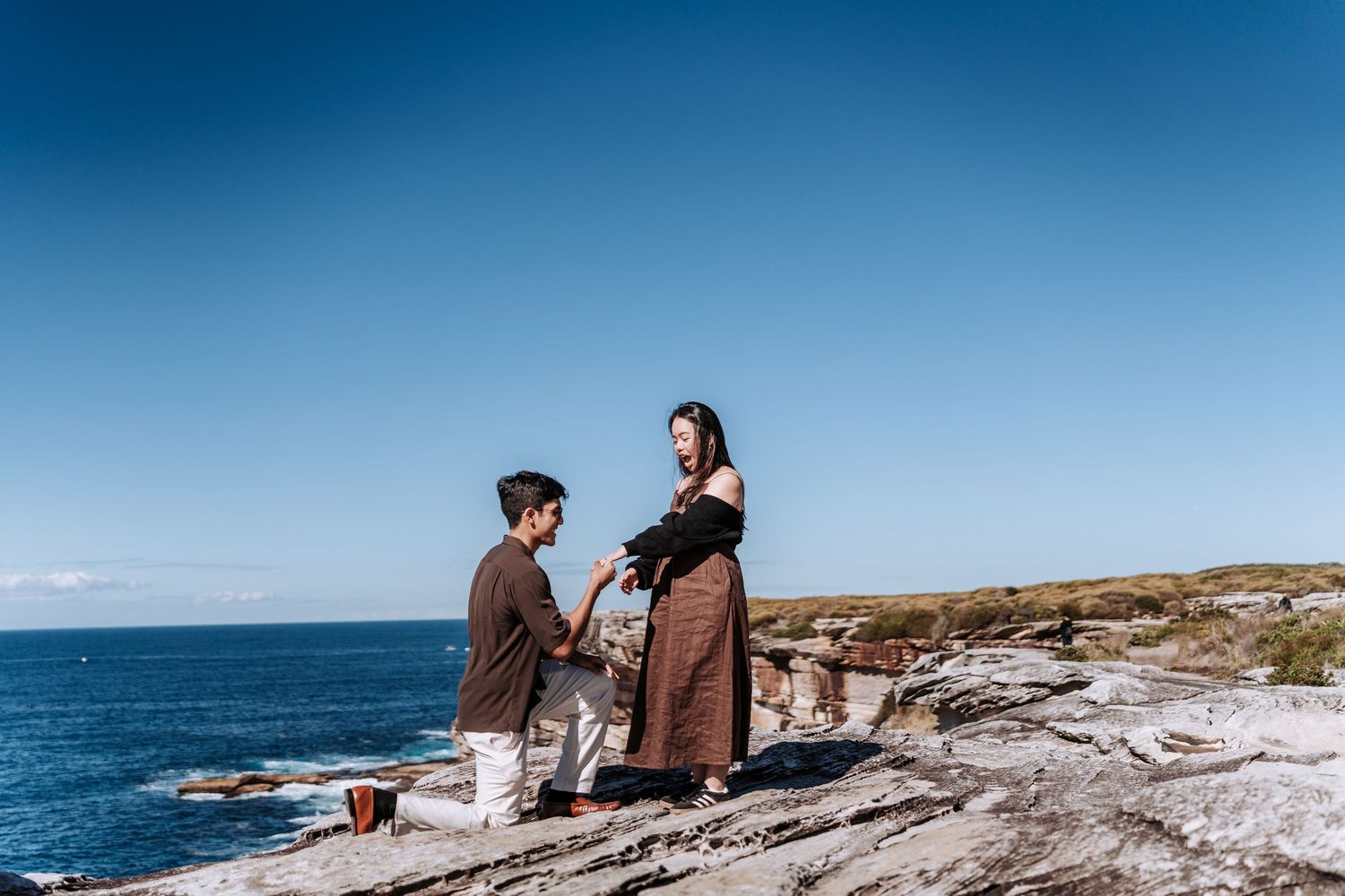 Man proposing to woman on rocky coastal cliff with ocean and blue sky backdrop.