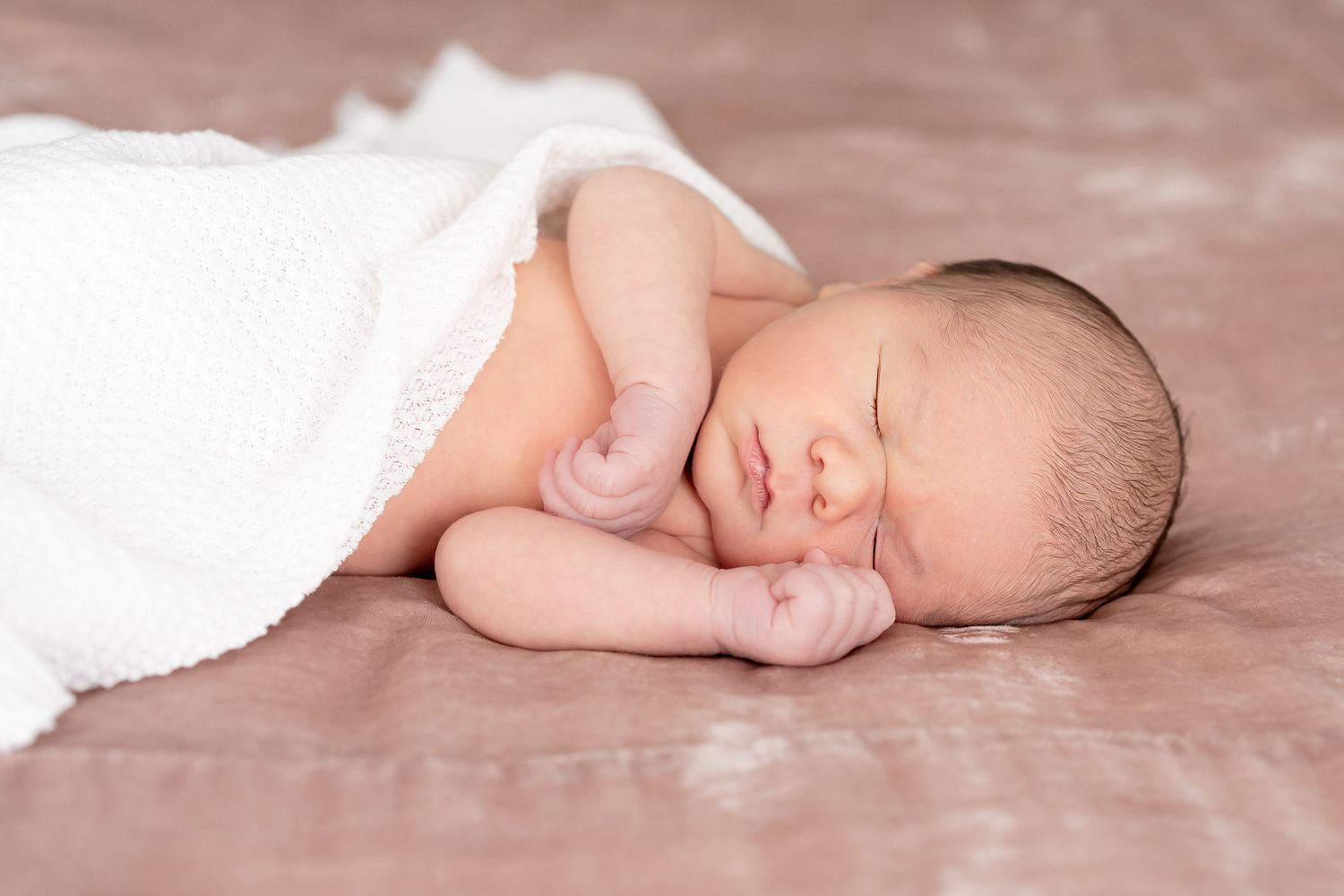 Sleeping newborn baby wrapped in white blanket lying on soft brown backdrop during professional photography session.