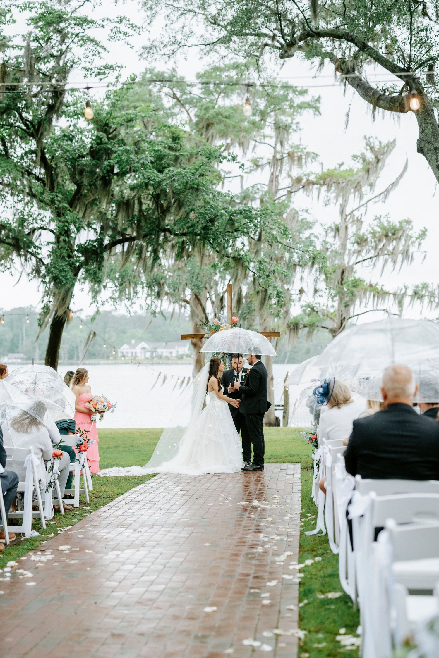 Bride and groom share vows under a clear umbrella at an elegant outdoor lakeside wedding ceremony with Spanish moss trees.
