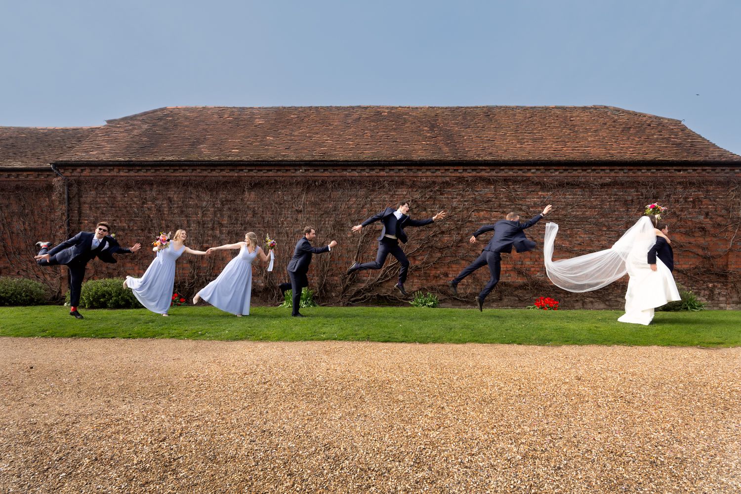 Wedding party joyfully running across green lawn holding hands, bride in white gown with flowing veil, beside rustic brick building.