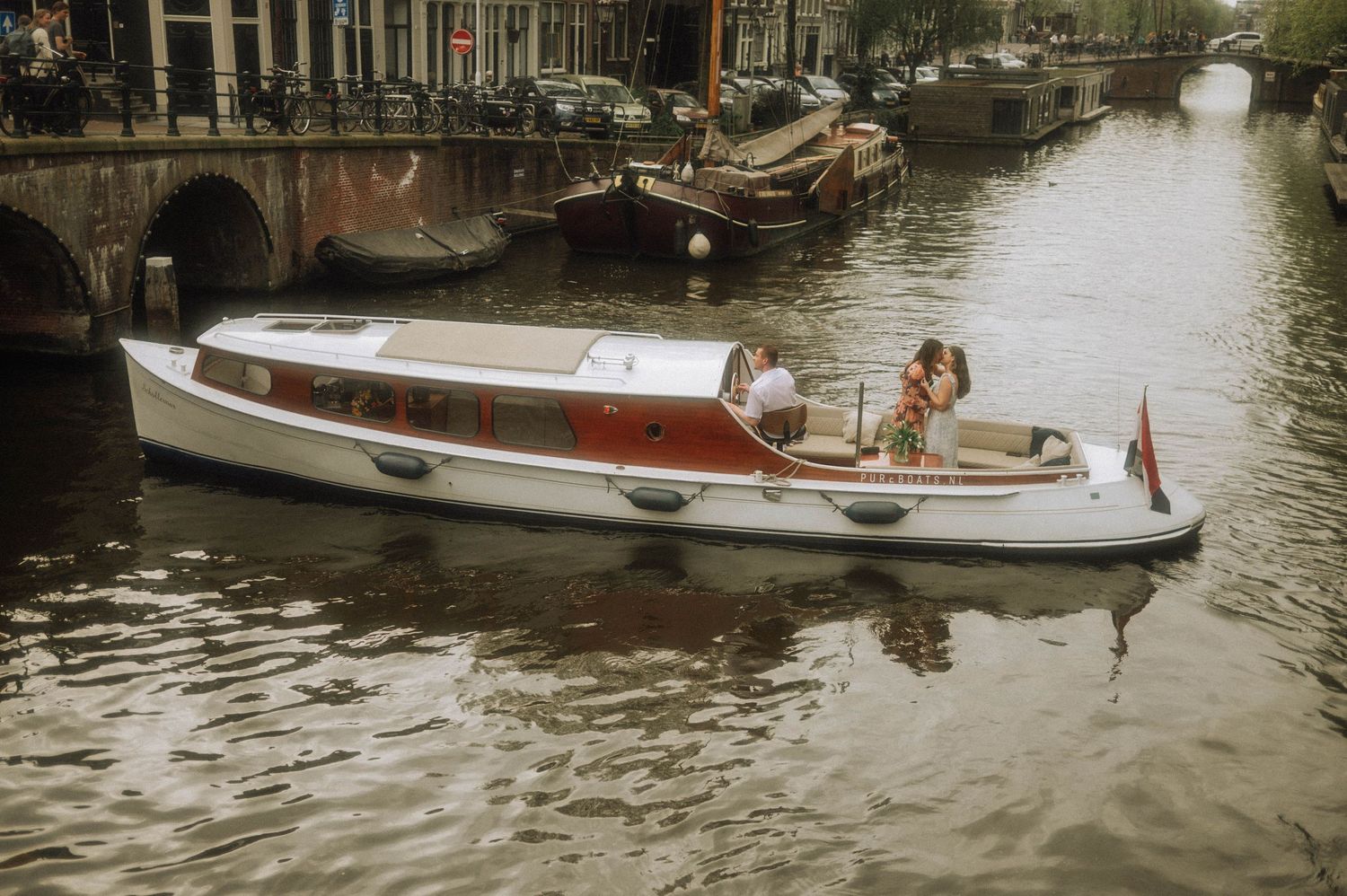 A white motorboat cruises along an Amsterdam canal with passengers aboard, historic buildings and moored boats in background.