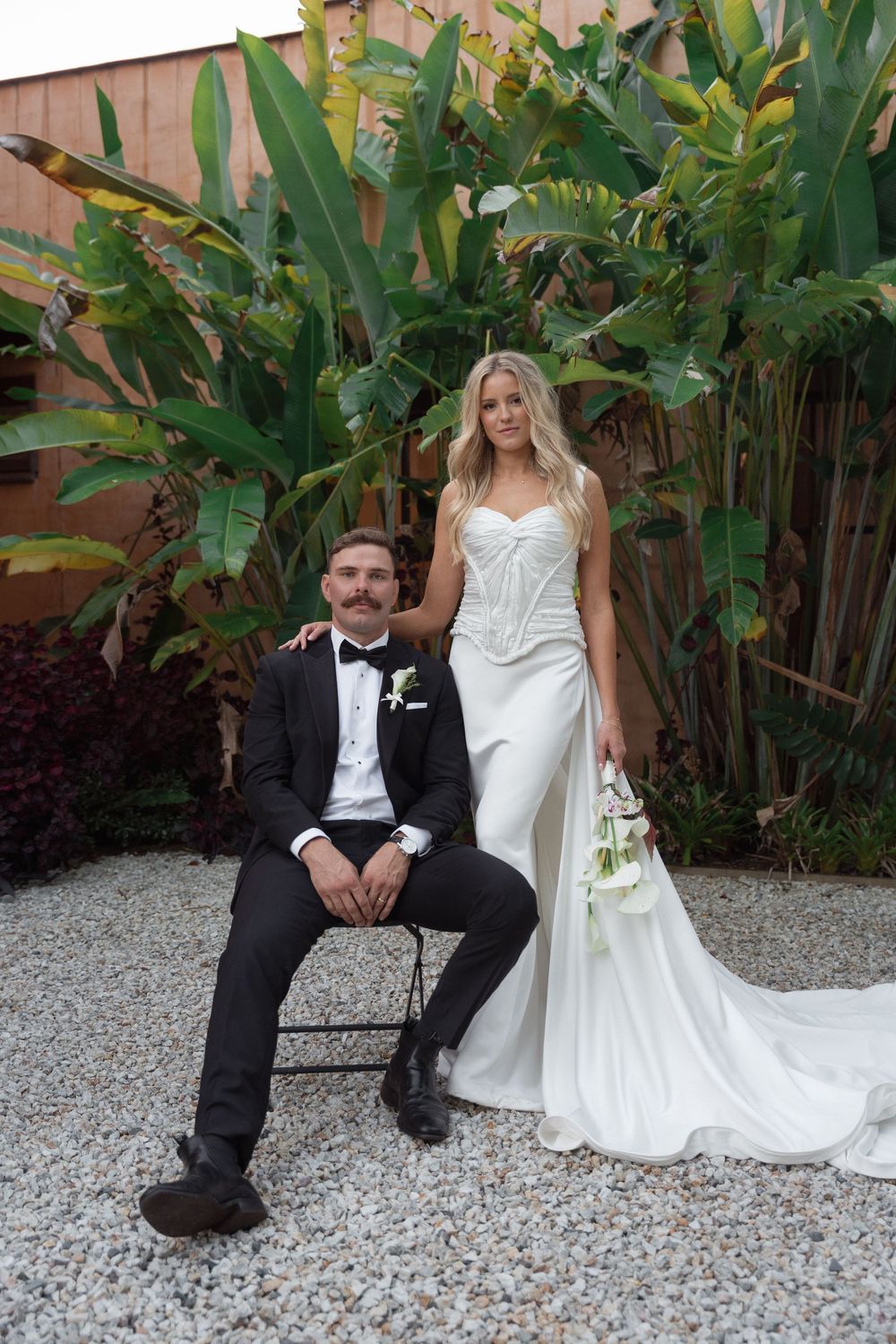Bride in white gown holds bouquet beside groom in black tuxedo seated on chair, with tropical greenery backdrop.