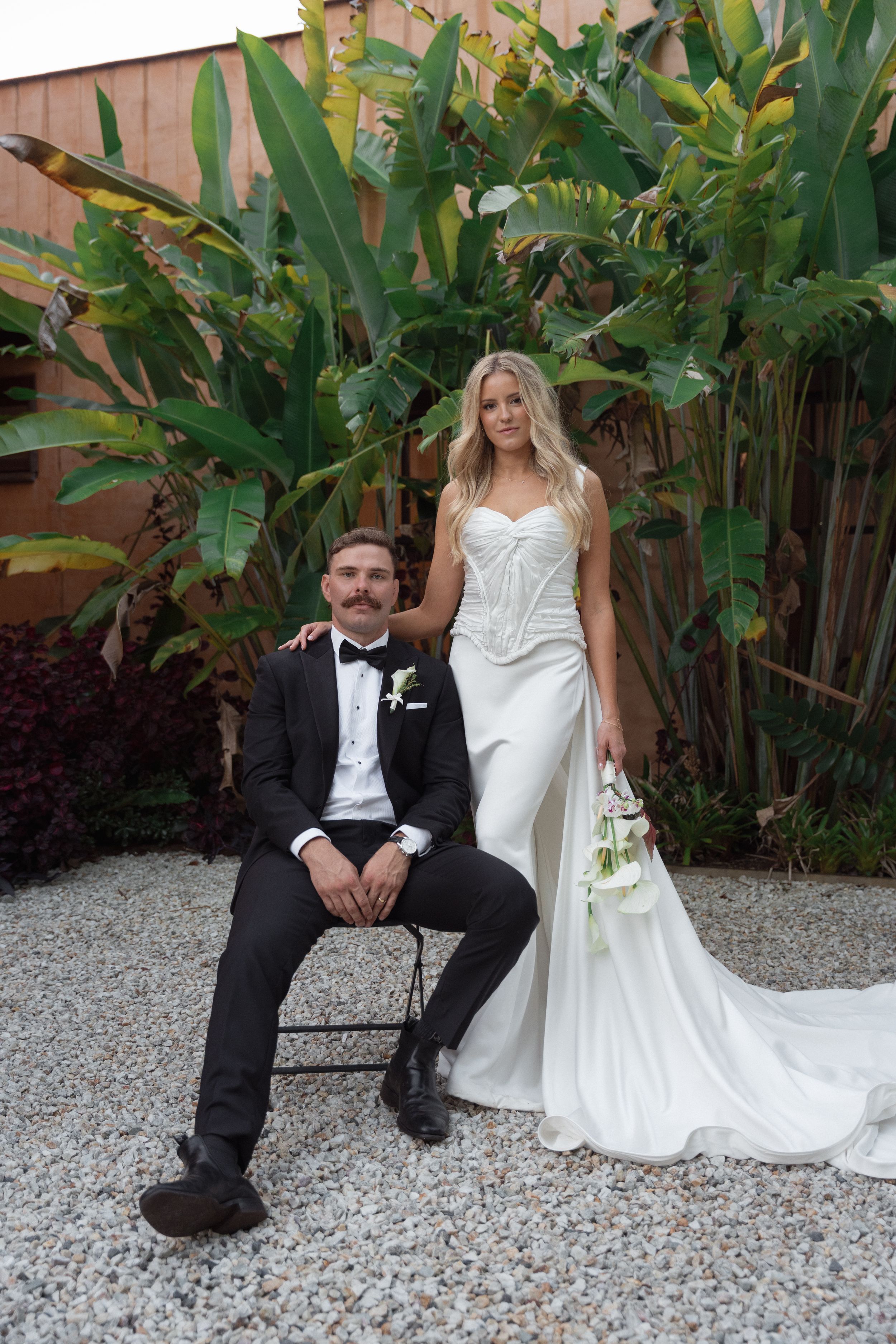 Bride in white gown holds bouquet beside groom in black tuxedo seated on chair, with tropical greenery backdrop.