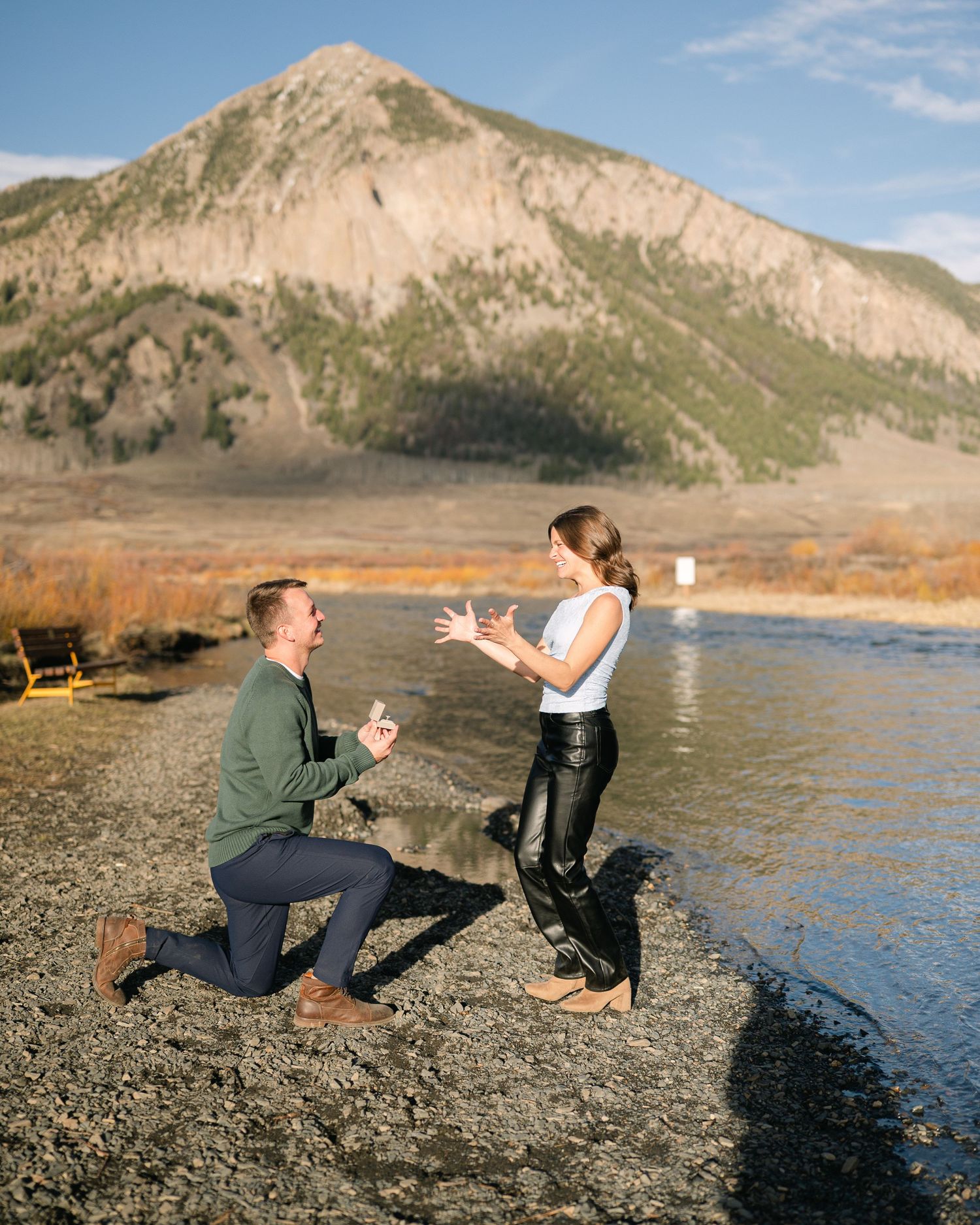 Happy couple laughing outdoors, man giving woman a piggyback ride in a scenic field with mountains in background.