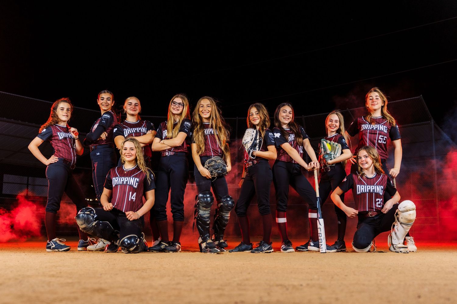Youth girls softball team posing in maroon uniforms with bats and gear against dramatic red smoke backdrop at night.