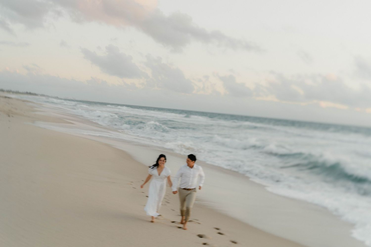 Two people in white attire walk hand in hand along a scenic beach shoreline at sunset with waves crashing nearby.