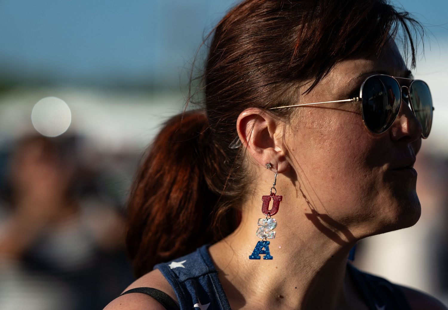 Woman wearing University of Utah and letter A sports fan earrings with aviator sunglasses at outdoor event.