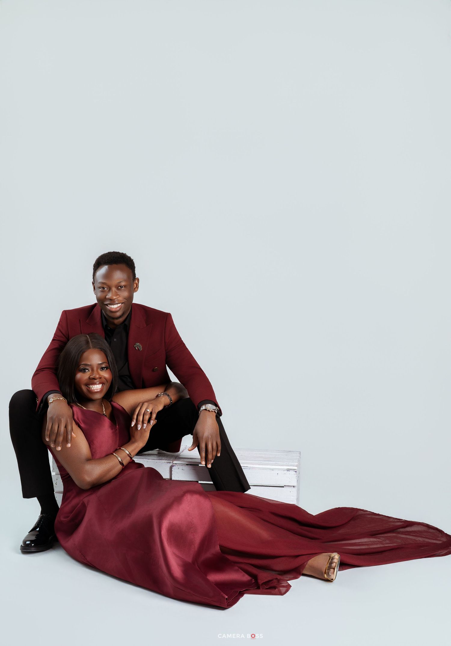 Couple in matching burgundy outfits posing elegantly in a studio, man in suit sitting behind woman in satin gown.