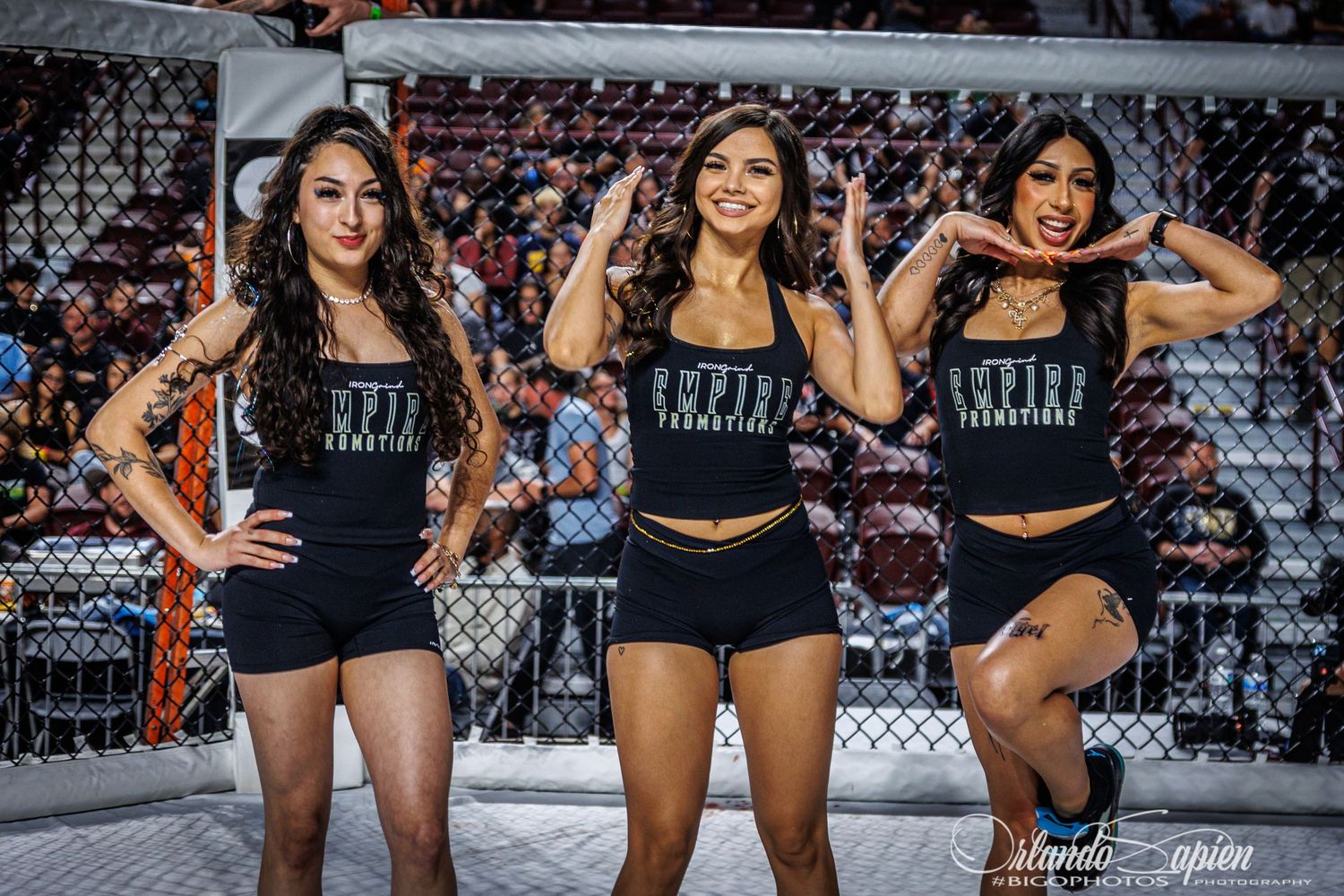 Three ring card girls in black outfits pose together in front of an MMA octagon cage during an event.