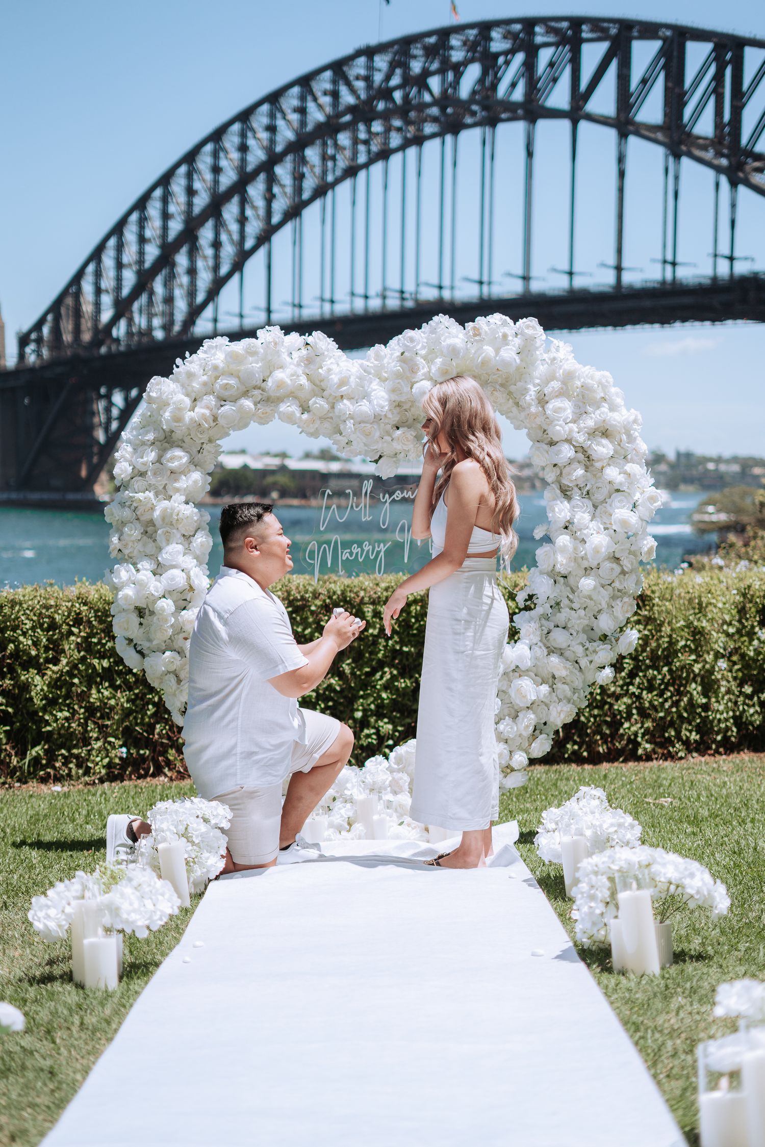 Couple exchanging vows at outdoor wedding ceremony with white floral arch, photographer capturing moment near Sydney Harbour Bridge.