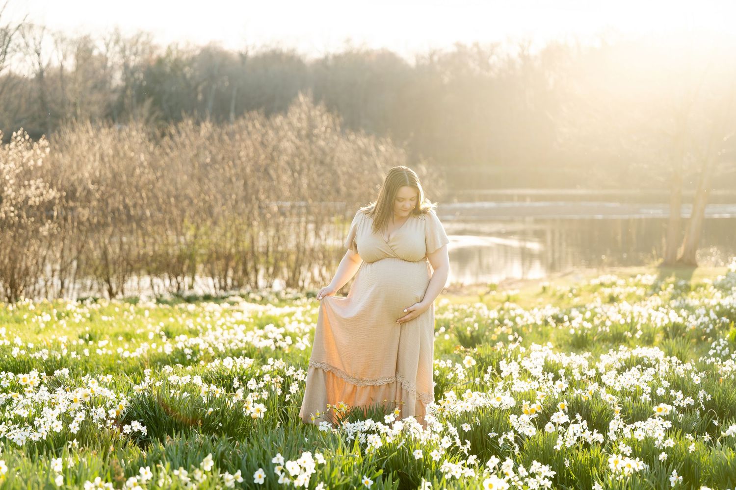 Pregnant woman in flowing pink dress standing in field of white daffodils during golden hour.