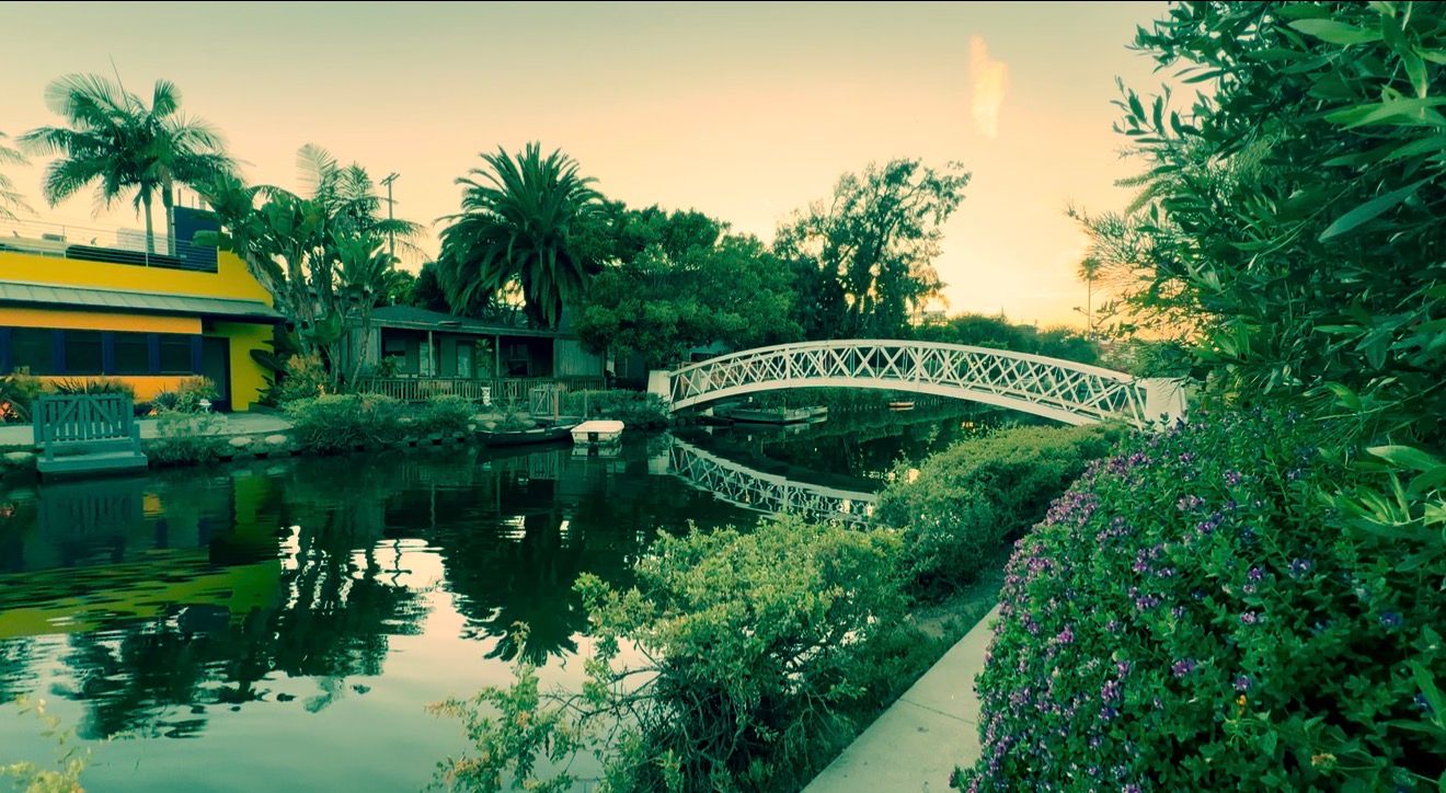 Scenic Venice Canal with ornate white footbridge, lush greenery, and golden sunset reflections in Los Angeles, California.