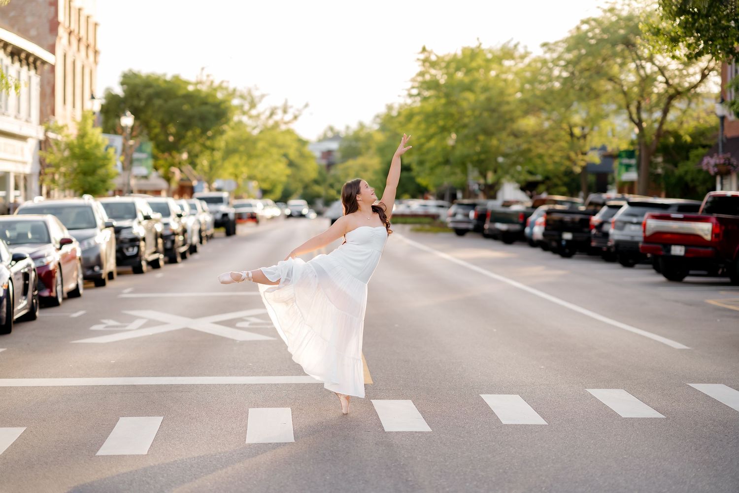 Ballerina in white dress dancing gracefully on a crosswalk in a sunny urban street with parked cars and trees.