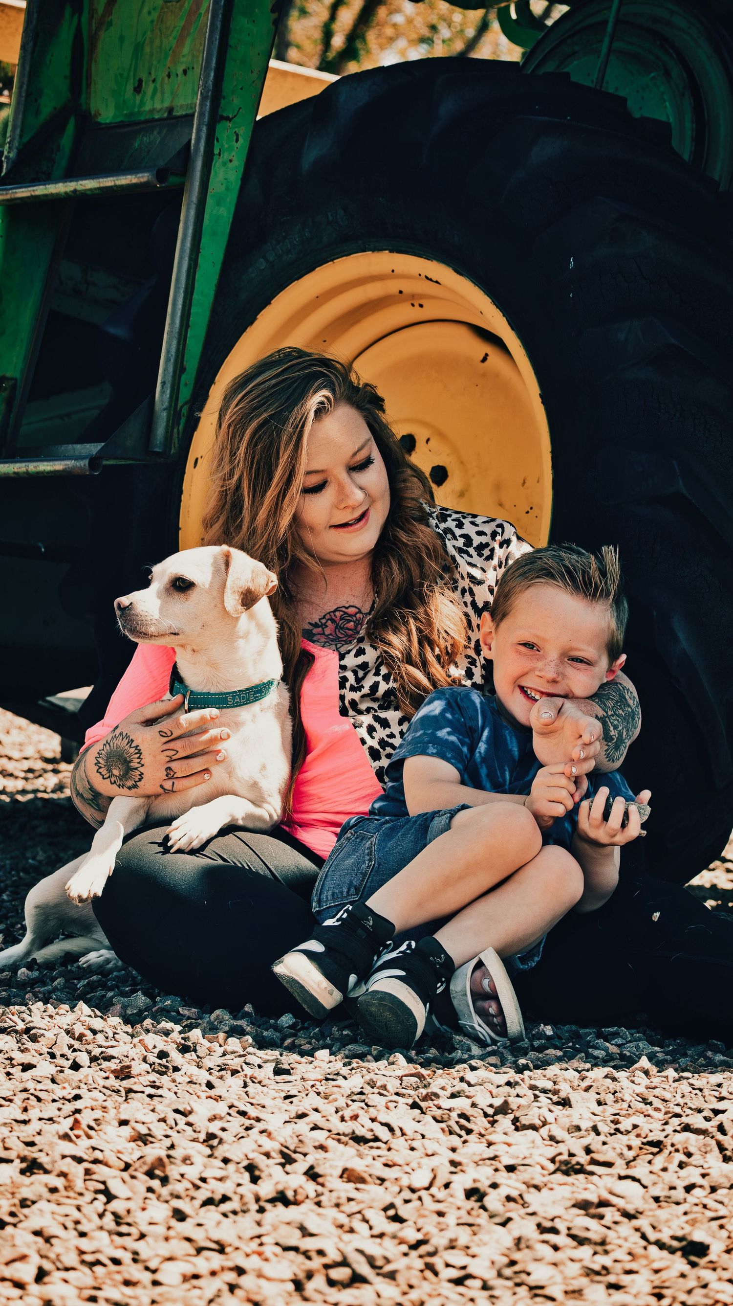 Woman and child sit laughing with a white dog in front of a green tractor tire on gravel.