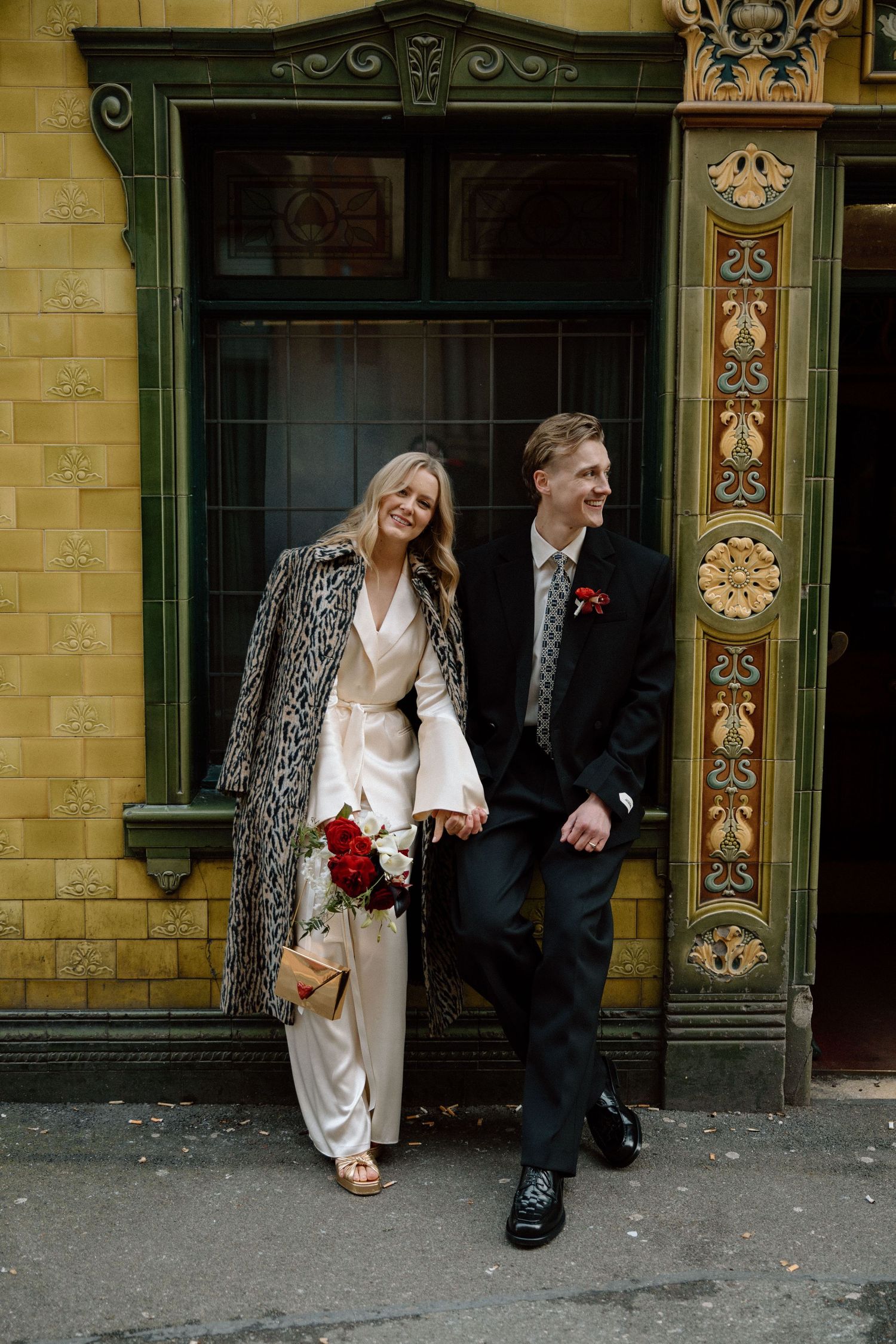 A smiling couple in wedding attire pose outside Peveril of the Peak pub in Manchester, she in a leopard coat and he in a dark suit.
