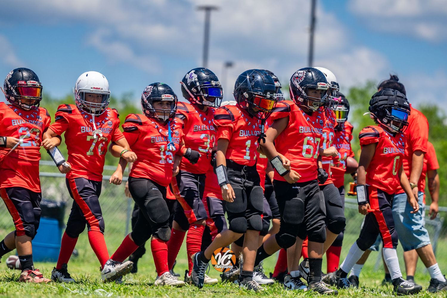 Youth football players in red uniforms and helmets walk together on a sunny field during a game day.