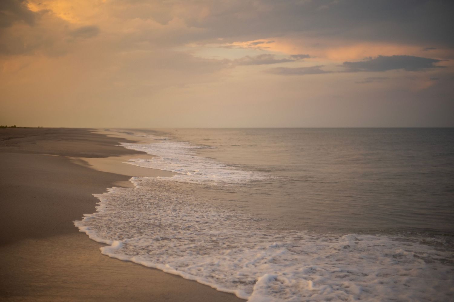 Serene beach at sunset with gentle ocean waves washing ashore on empty sandy coastline under golden cloudy sky.