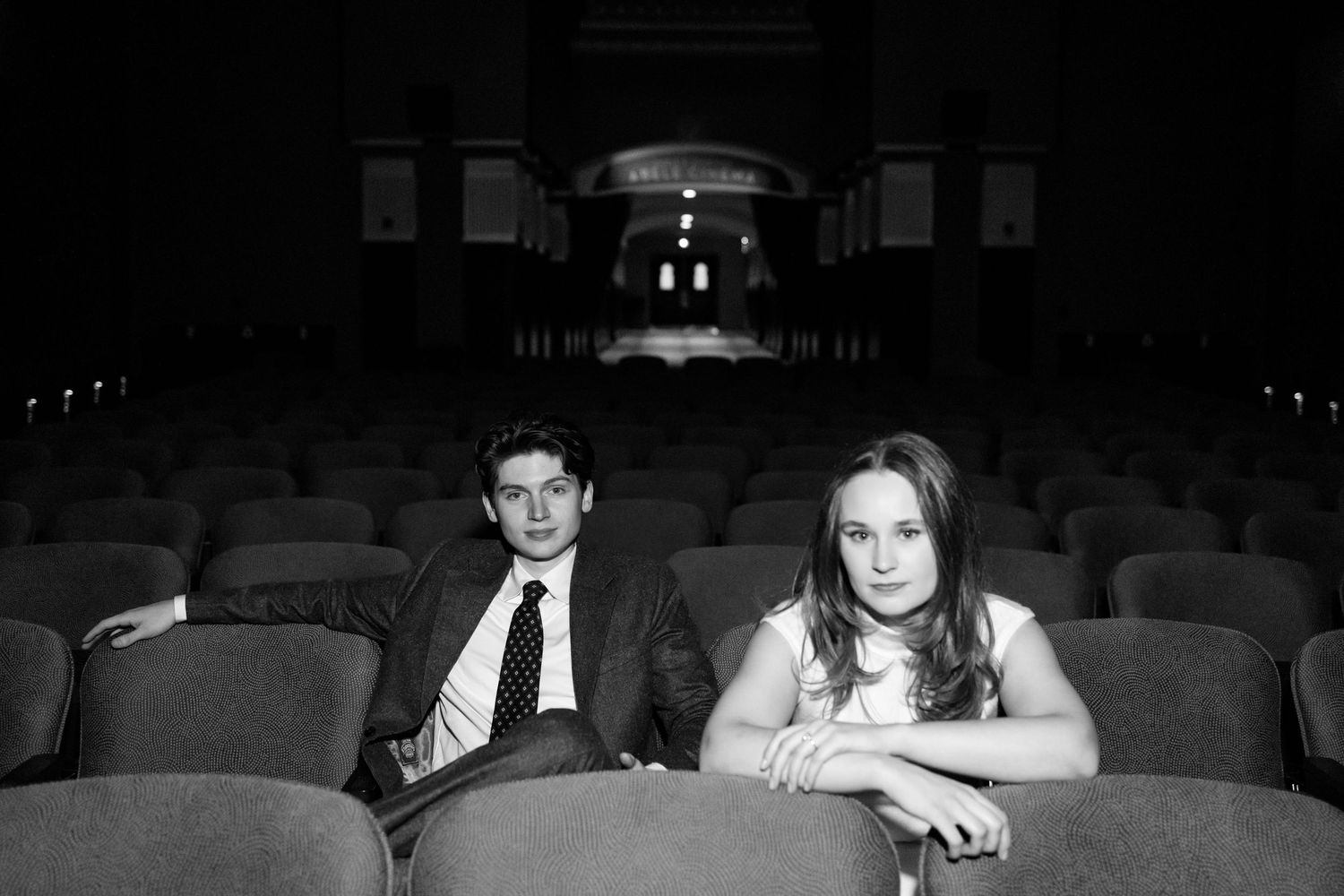 Black and white photo of a young man and woman sitting in empty theater seats, looking contemplative.