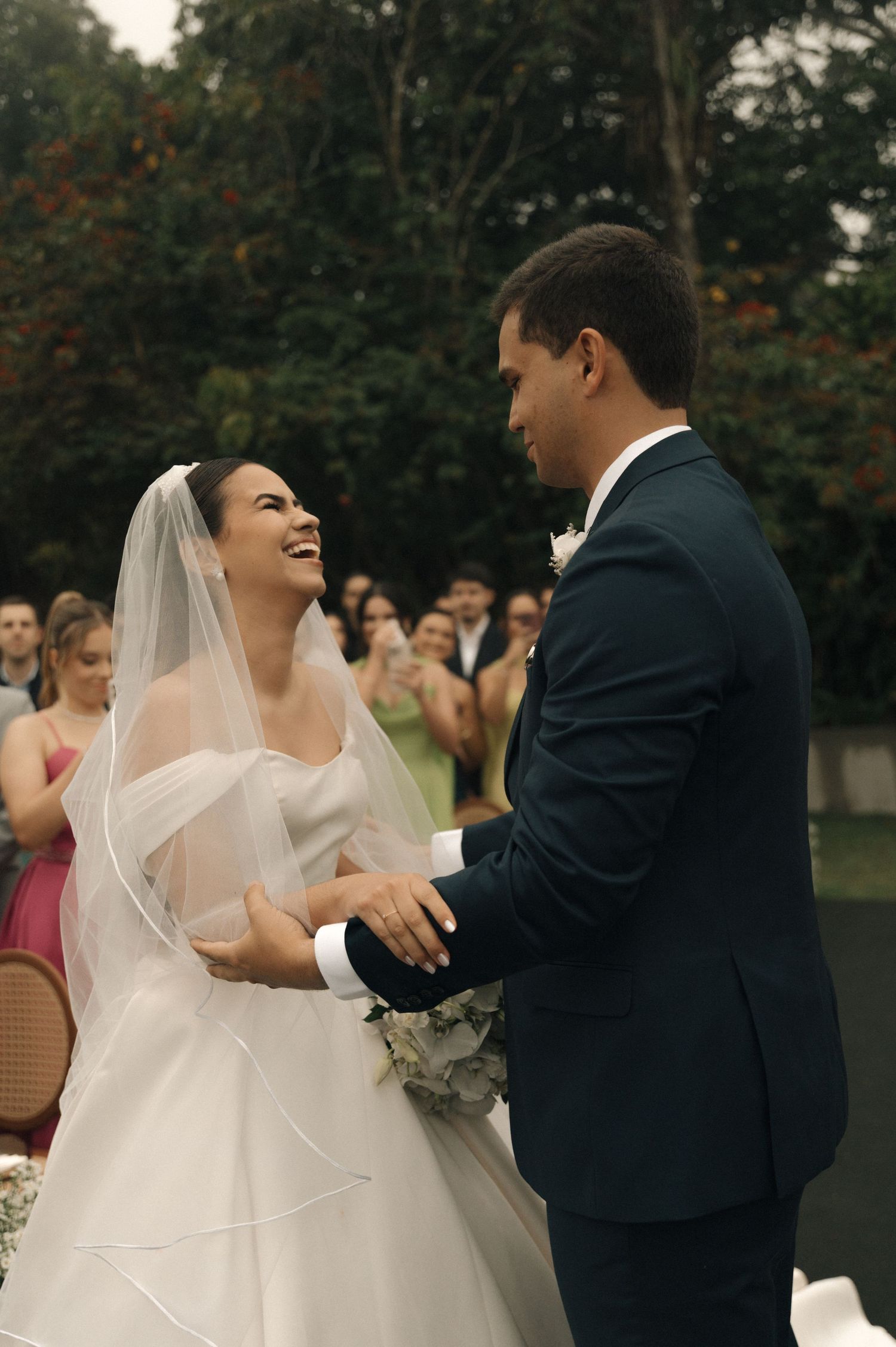 Bride in white gown and veil laughing joyfully with groom in navy suit during outdoor wedding ceremony.