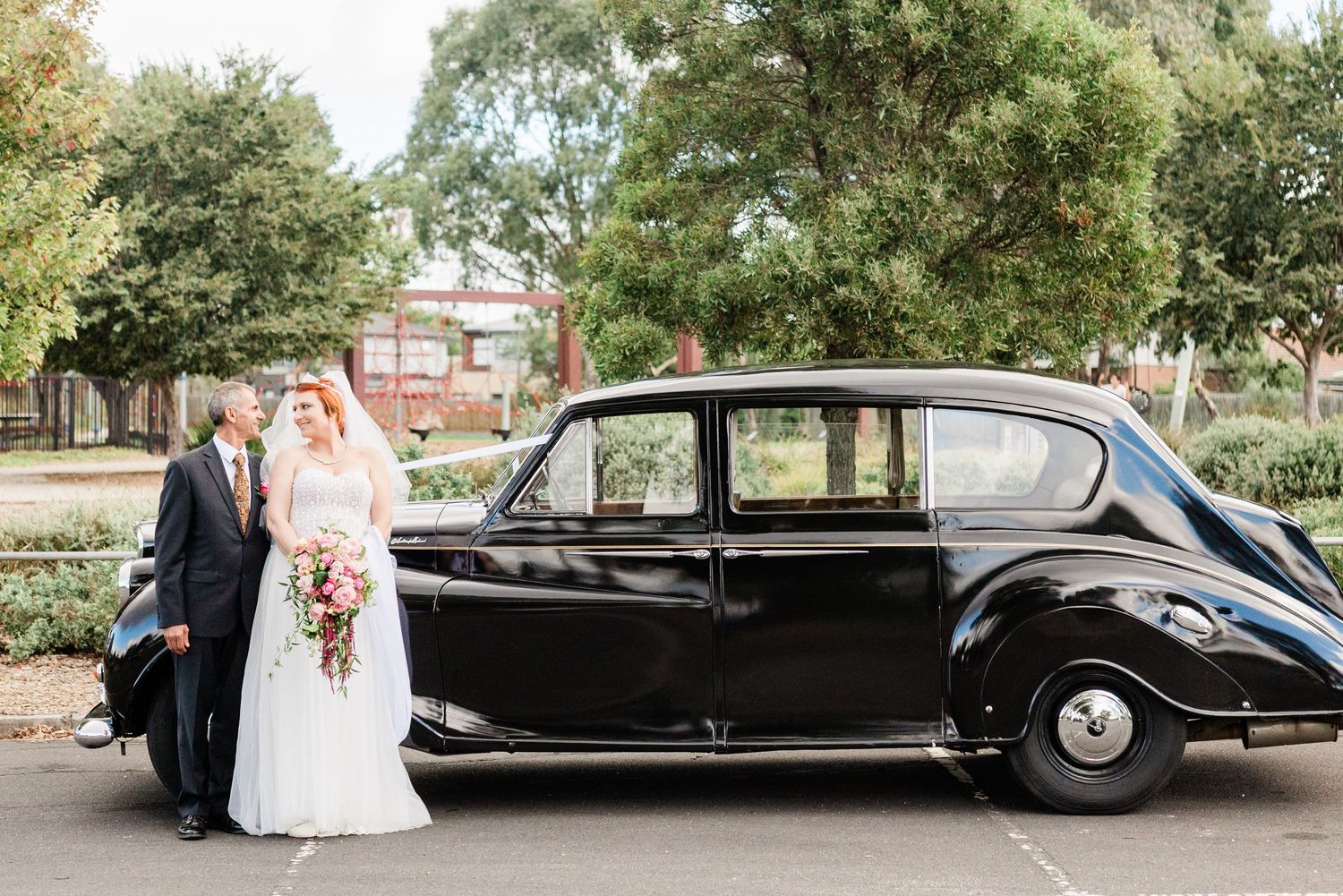 Bride and groom posing beside a classic black vintage car outdoors on their wedding day.