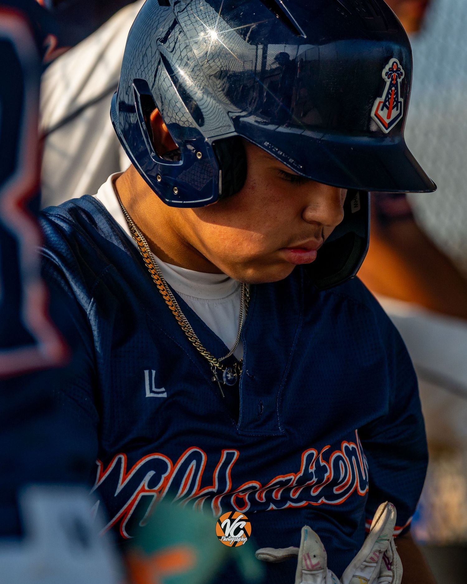 Baseball player in navy helmet and jersey with gold chain looks down in dugout during game.