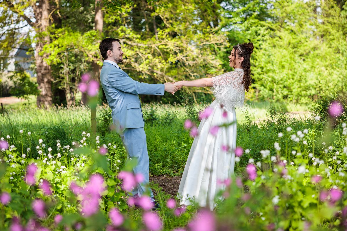 A bride and groom hold hands dancing in a lush green garden with blooming pink and white wildflowers.