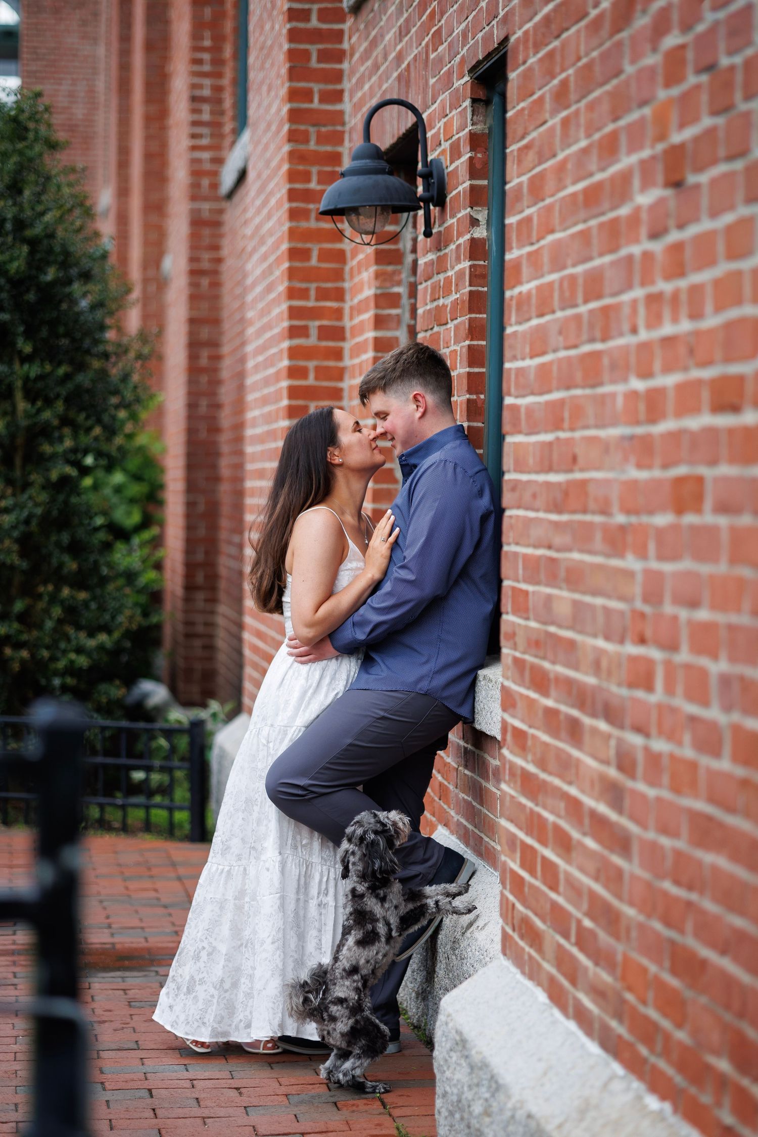 Couple in wedding attire with small dog on cobblestone street between historic brick buildings with green bridge overhead.