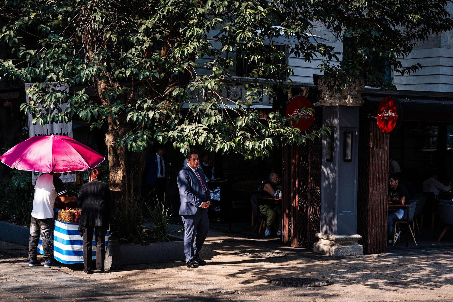Pink umbrella provides shade on a sunny sidewalk near storefronts and trees in an urban setting.
