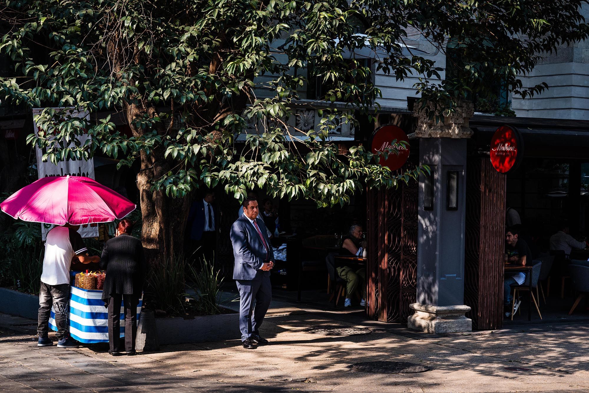 Pink umbrella provides shade on a sunny sidewalk near storefronts and trees in an urban setting.