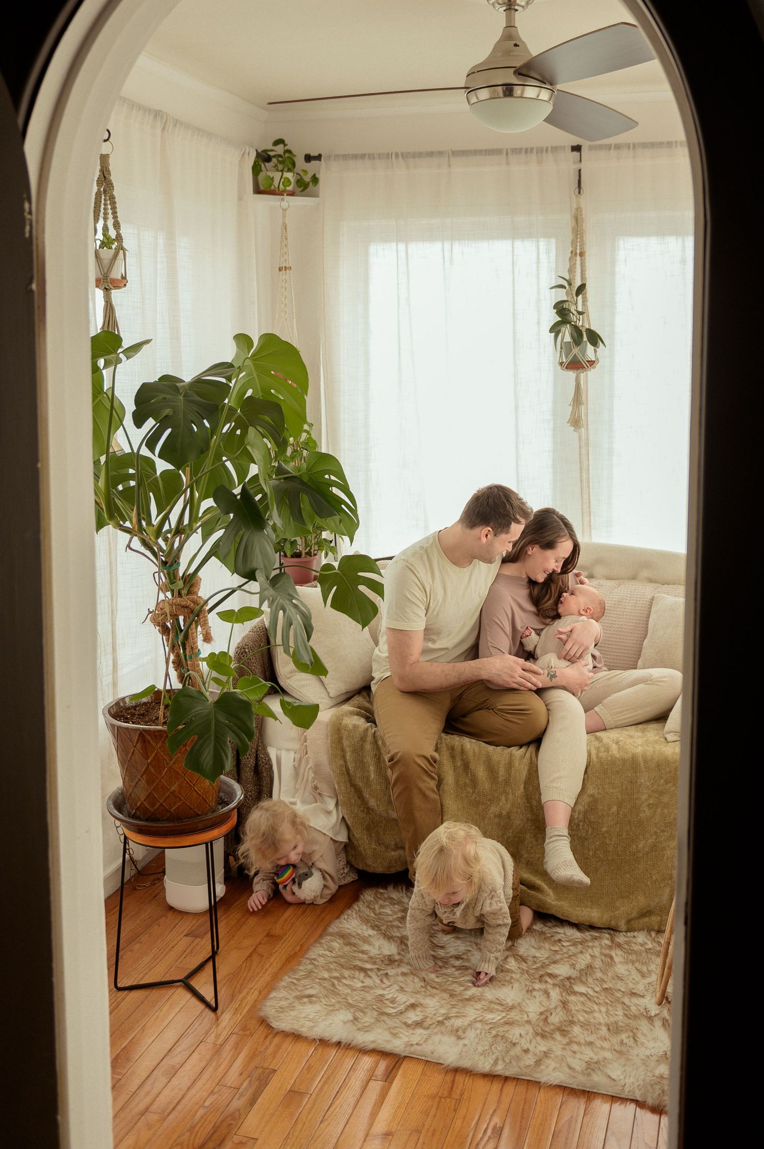 Family with newborn baby sitting on couch while toddlers play on floor in bright living room with plants.