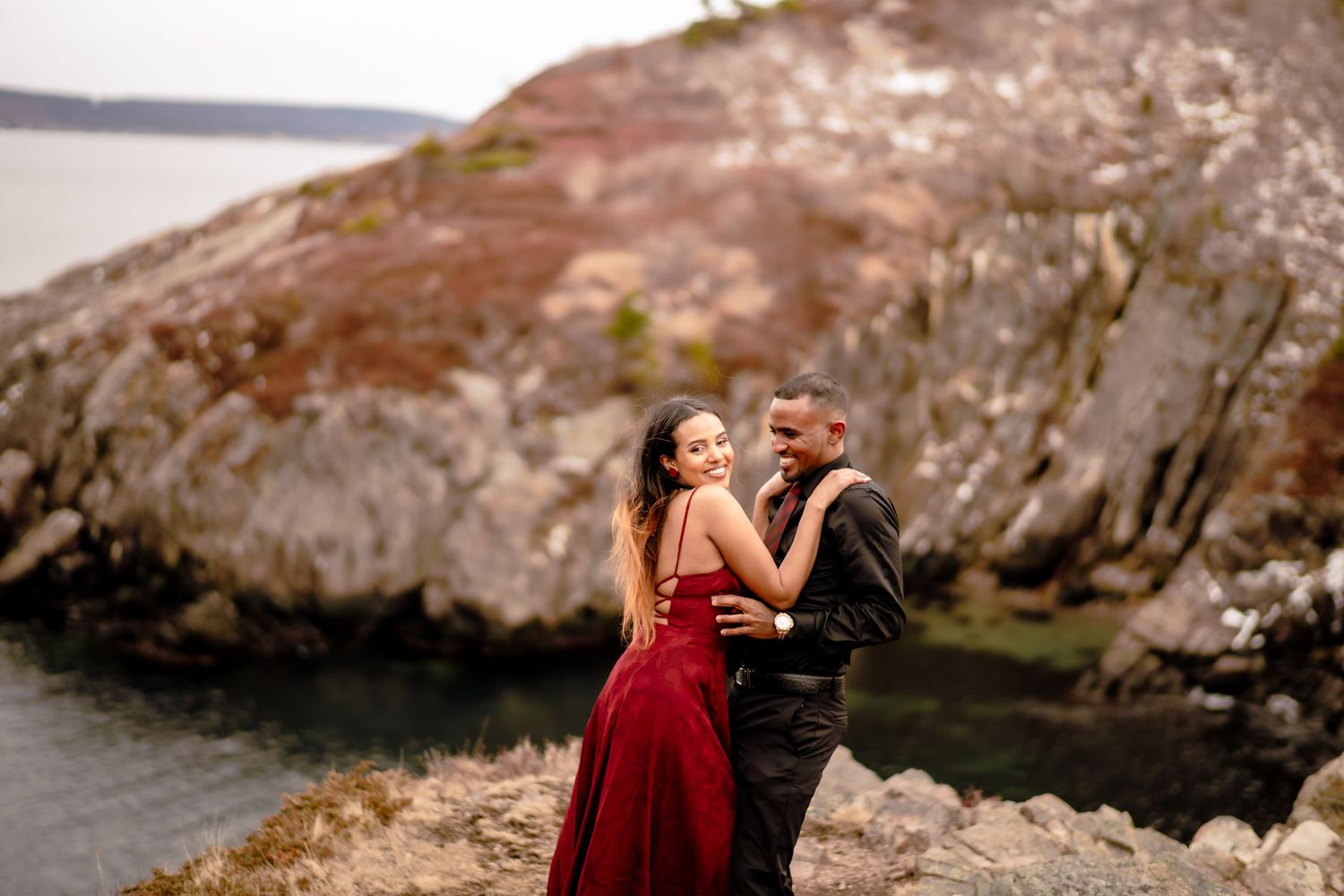 couple embracing with ocean view in the background, woman in red dress and man in black, St. John's Newfoundland engagement photography