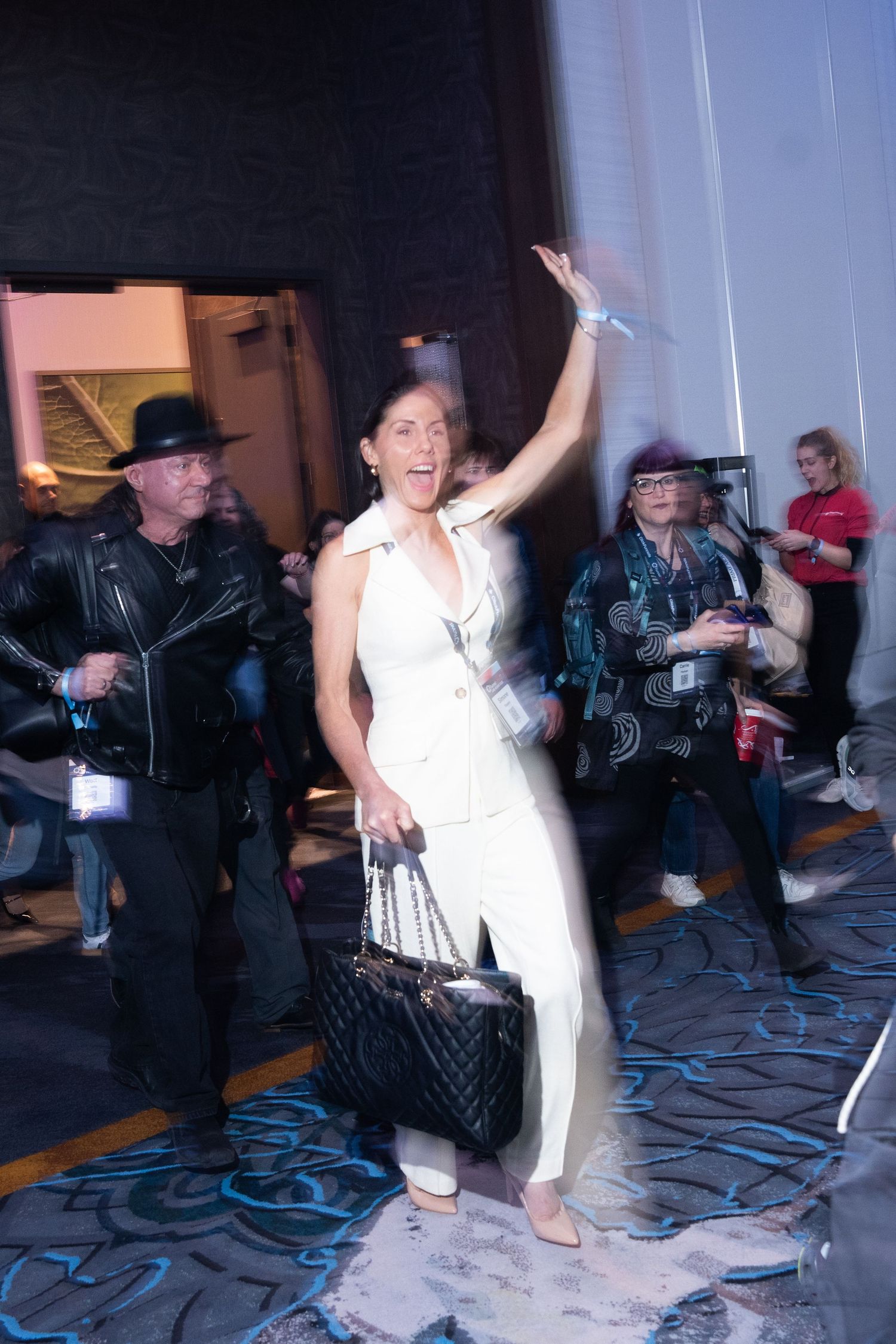 Woman in white suit waving at event, holding quilted black bag, surrounded by crowd in dimly lit venue blurred with a creative shutter drag