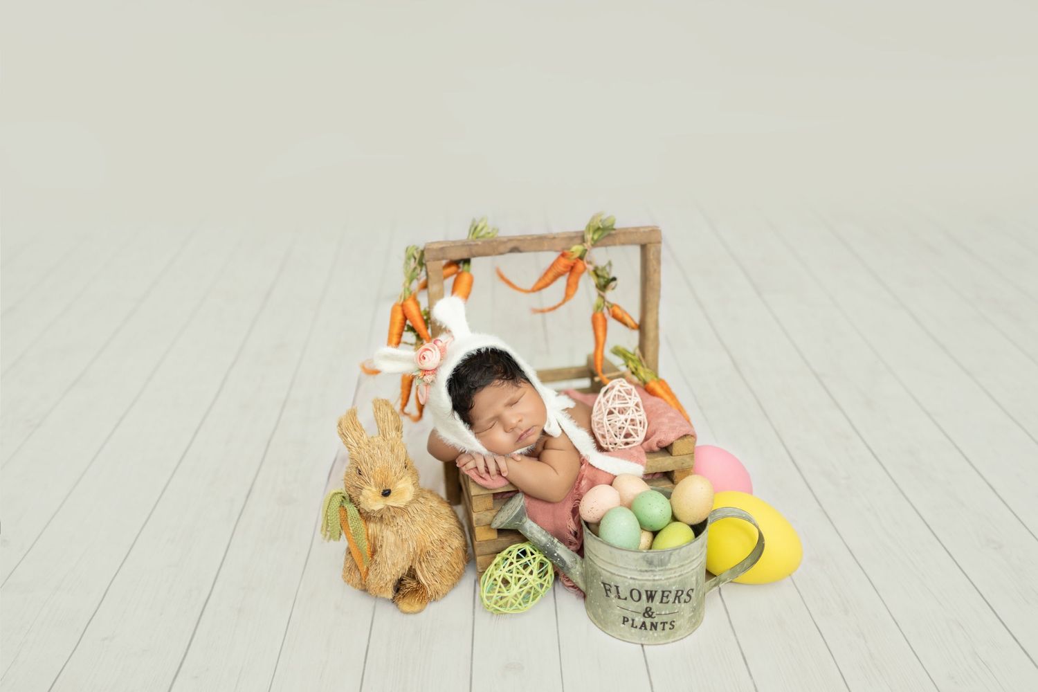 Newborn baby sleeping in Easter-themed photo with bunny, colorful eggs, carrots, and flower bucket on white wood floor.