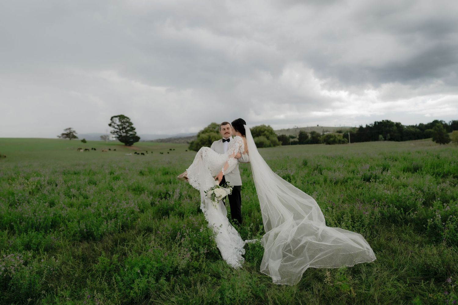 Bride and groom embrace in a lush green field, her veil flowing dramatically in the wind under a stormy sky.