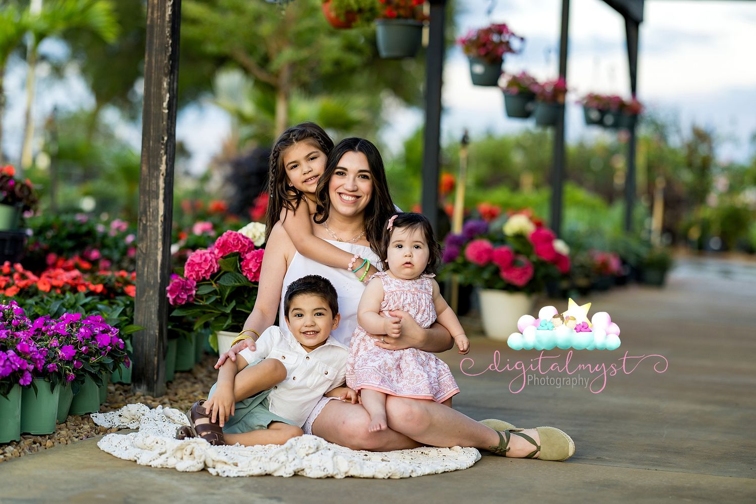 A mother poses with her three young children among colorful flowers at an outdoor garden center.