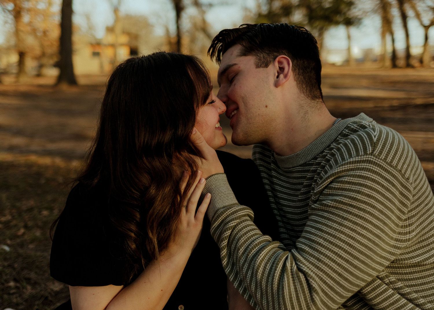 A couple shares a tender almost-kiss moment outdoors in a park during golden hour.