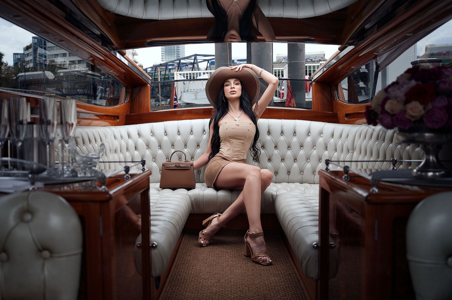 Elegant woman in beige dress and hat poses on luxury yacht interior with white tufted seating and designer handbag. Shot by Boyd Visuals.