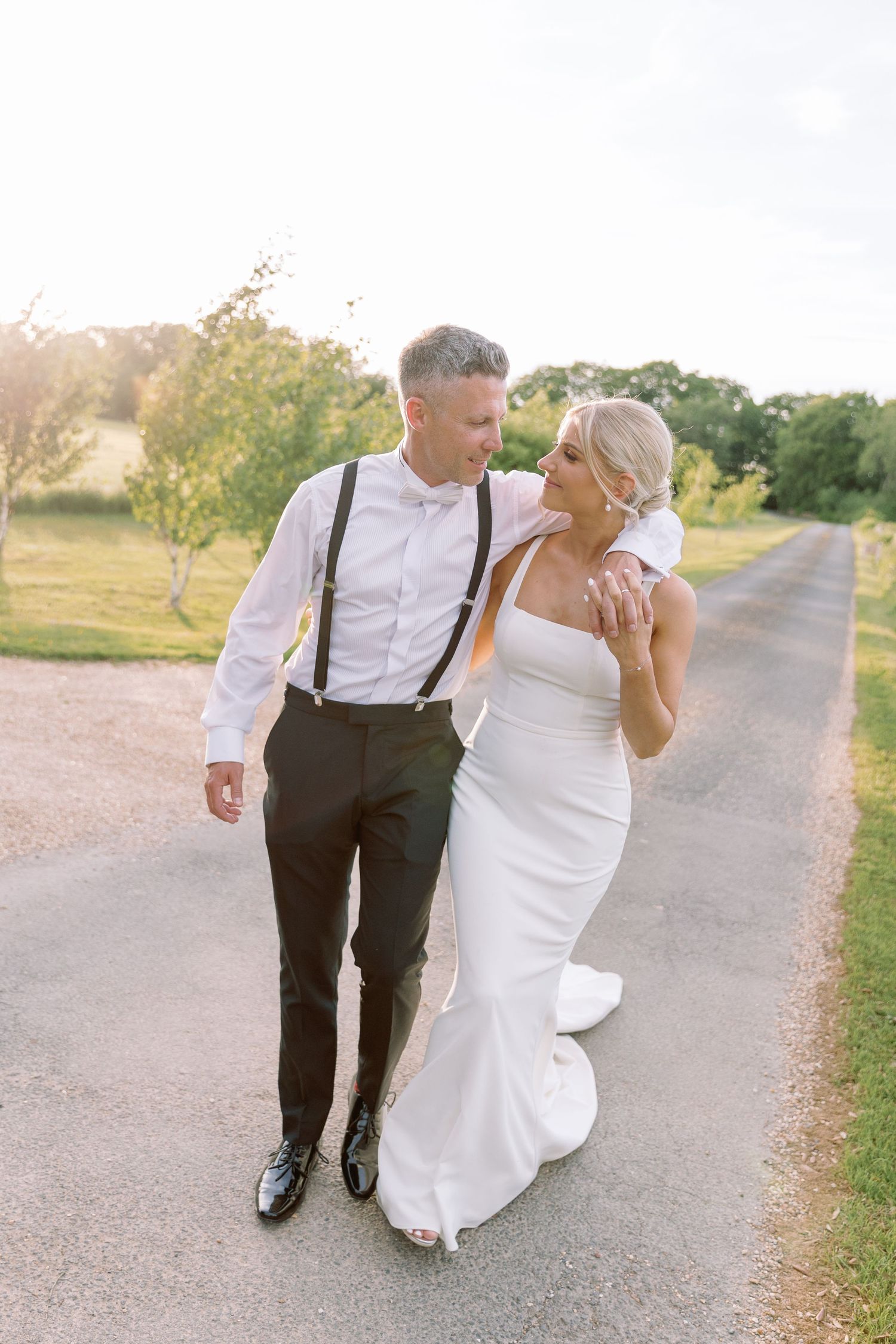A couple in wedding attire walks and laughs together on a country path during golden hour.