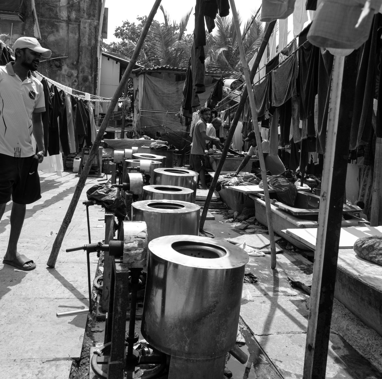 Black and white image of a man standing beside large metal cylinders in an outdoor urban workshop setting.
