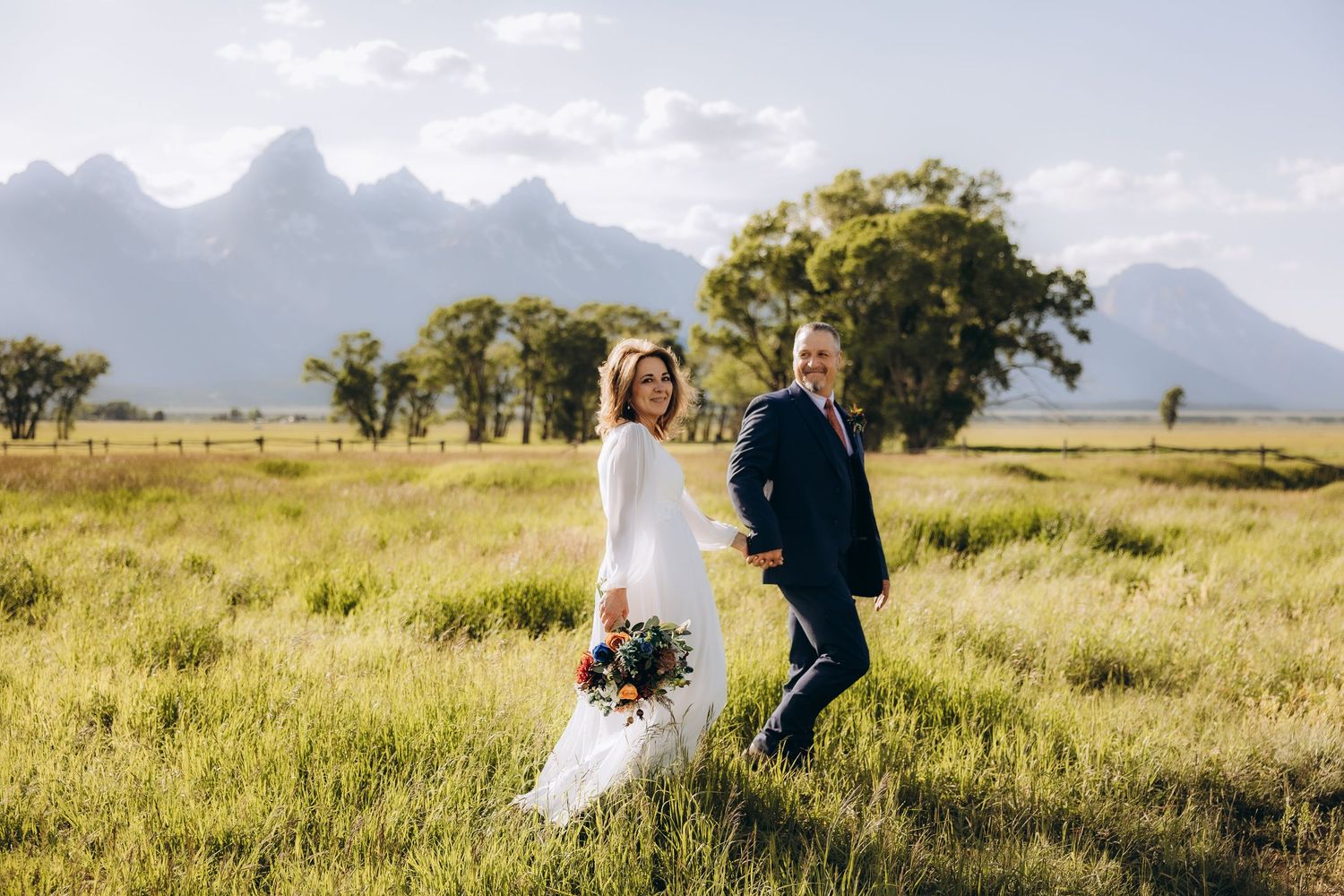 A couple in wedding attire walks through a scenic golden field with mountains and trees in the background.