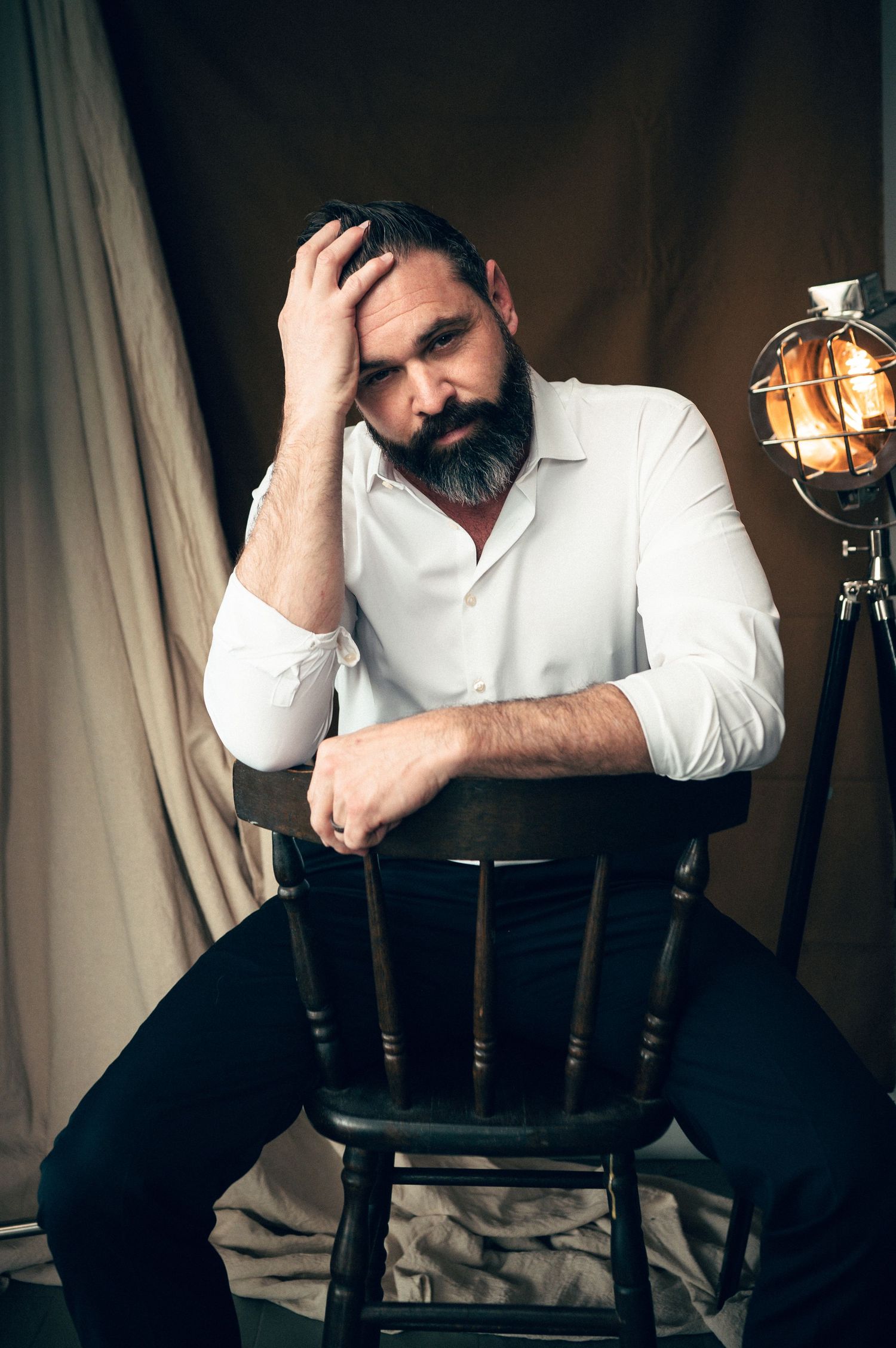 Salt lake city Utah Headshot portrait by Carl James Photography. Bearded man in white shirt sitting on wooden chair with hand in hair.